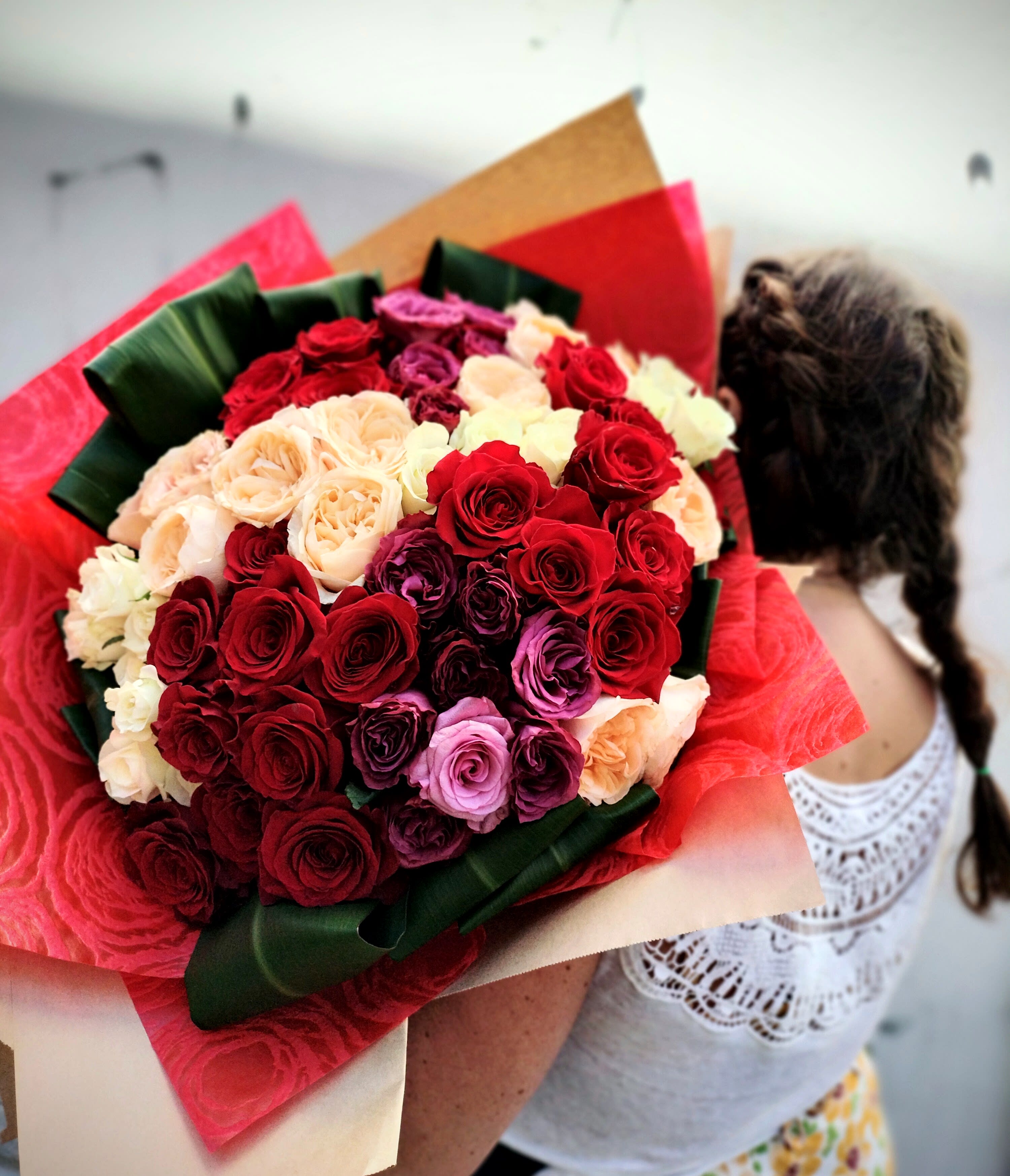 Large hand-tied bouquet of red, cream, and purple roses wrapped in red and kraft paper, held by a person.