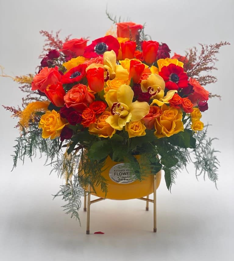 Morning Glory - The contrasting colors of yellow, orange, and red in this arrangement create a dynamic visual display, with the warmth of the colors evoking a sense of energy and excitement. The arrangement is carefully balanced, with the taller flowers at the back of the vase and the smaller flowers towards the front, creating a natural flow and sense of movement. The yellow and orange spray roses add texture and dimension, with their delicate petals and intricate centers. The metallic stand provides a sleek and modern touch, while the yellow ceramic vase ties the whole arrangement together, complementing the colors of the flowers.  The combination of the gold metallic stand and the yellow ceramic vase creates a striking visual contrast, with the warm color of the vase offset by the metal of the stand. The height of the stand also elevates the arrangement, making it more noticeable and eye-catching.  This arrangement is named "Morning Glory" becasue it evokes a sense of freshness, energy, and new beginnings.  Overall, this flower arrangement is a beautiful and carefully crafted display of colors, textures, and shapes. It would be a perfect choice for anyone looking to add a touch of energy and vibrancy to their living space or special event.