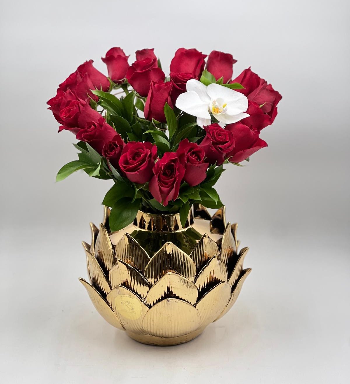 Red roses arranged in a gold lotus-shaped vase with one white orchid bloom
