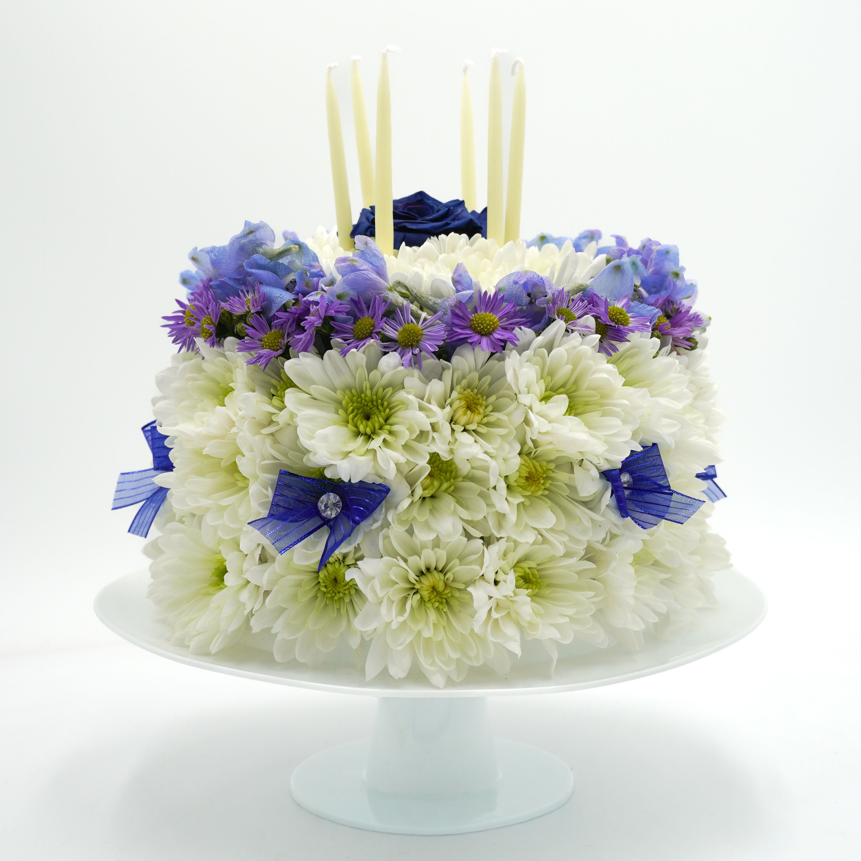 Blue Elegance Birthday Cake - A floral "cake" fashioned from dense white chrysanthemums with a halo of small purple daisy-like blooms and a single deep blue rose crowning the top-perfect as a birthday arrangement or a congratulatory centerpiece. Styled with tall ivory candles and cool-toned blues and purples, this birthday bouquet makes the recipient feel celebrated and seen, whether it's a milestone year or a special achievement.  The base is a compact mound of fresh white chrysanthemums, a middle ring of petite purple daisies, and accents of cream roses and blue ribbon woven around the sides for a tidy, elegant finish. A striking deep blue rose sits at the center top surrounded by several tall ivory candles on a white plate, giving the piece a formal, ceremonial silhouette ideal for table display.  Designed to read as a gift and a centerpiece, this arrangement works equally well for birthday tables or to say "congratulations" at an event. Measures to fit standard tabletops; keep candles attended and remove before placing in direct drafts. Order with same-day delivery available when freshness matters.