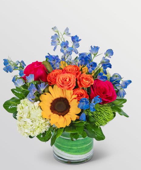 Havana Heat - Ignite joy and color with Havana Heat, a dazzling bouquet bursting with tropical energy and charm. This radiant arrangement features sunny golden sunflowers, fiery orange spray roses, bold hot pink roses, and sky-blue delphinium, all beautifully offset by crisp hydrangeas, unique green trick dianthus, and glossy greens.  Expertly arranged in a chic glass vase wrapped with a fresh green ribbon detail, this bouquet is your go-to choice for birthdays, celebrations, thank-you gifts, or to instantly brighten someone's day.  Features: Vibrant mix of sunflowers, roses, delphinium, and more Artfully designed in a compact round glass vase (included) Bold, tropical-inspired color palette Note: Flower availability may vary slightly, but substitutions will maintain the bouquet’s joyful and colorful essence.