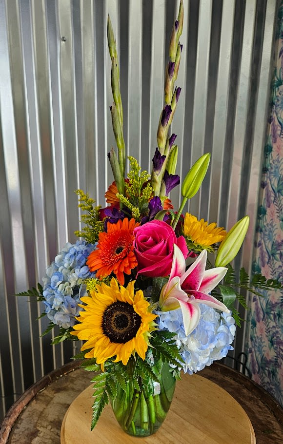 Summer Carnival - A festival of flowers for every joyful occasion. Bursting with bold color and vibrant personality, our Summer Carnival bouquet is a joyful celebration in bloom. This eye-catching arrangement features radiant sunflowers, vivid pink roses, stargazer lilies, orange gerbera daisies, deep purple gladiolus, and sky-blue hydrangeas — a kaleidoscope of summer’s finest. Presented in a clear glass vase, this design is perfect for birthdays, congratulations, or any moment that calls for bold beauty and big smiles. Highlights: Includes sunflowers, roses, stargazer lilies, gerbera daisies, gladiolus, hydrangeas, and assorted greenery Handcrafted with premium blooms for maximum impact