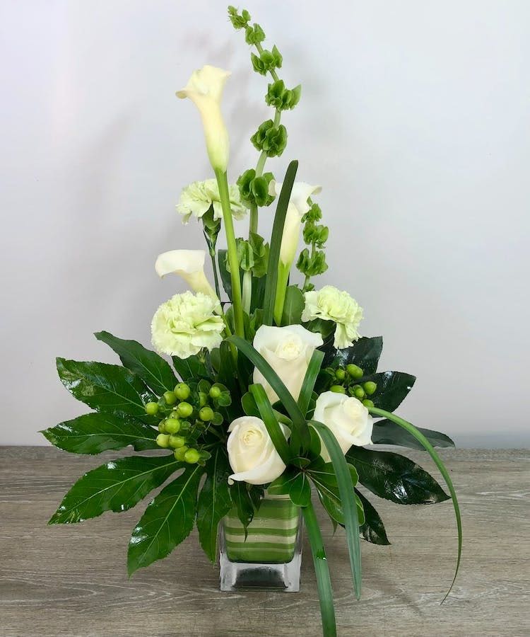 Artful Greens  - Sophisticated, modern, and beautifully sculptural, this elegant green and white floral arrangement is designed with clean lines and contemporary garden styling. Featuring premium white roses, graceful calla lilies, Bells of Ireland, green hypericum berries, and fresh tropical foliage, this upscale floral design offers a refined natural look with artistic movement and texture. Designed in a sleek glass cube vase with layered greenery accents, this arrangement is perfect for sympathy, corporate gifting, modern home décor, thank you gifts, anniversaries, or elegant everyday occasions. Handcrafted by the experienced floral designers at Purple Orchid Flowers and available for flower delivery throughout the Omaha metro area.