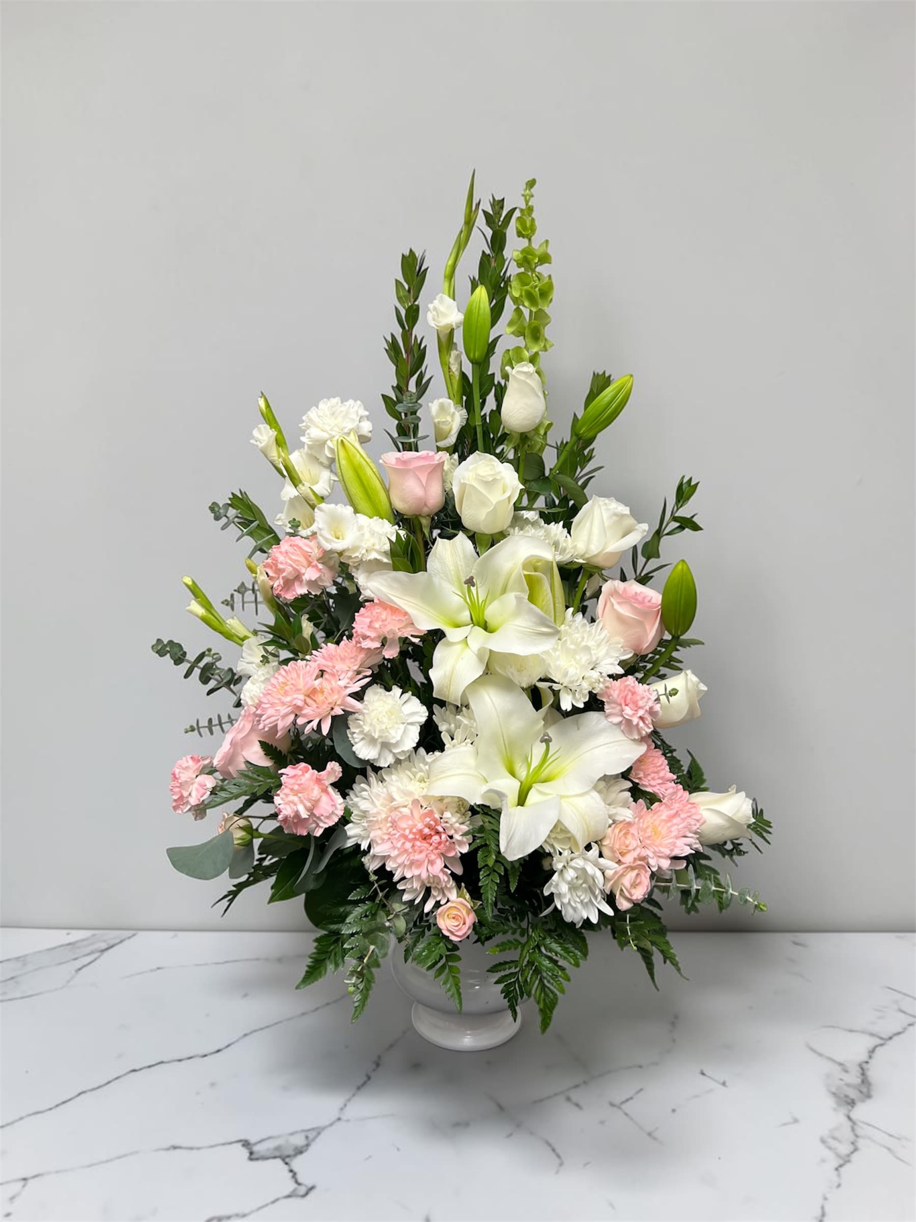 Pink and White Sympathy Arrangement  - A soft, layered sympathy arrangement that reads as both peaceful and gently romantic, perfect for expressing condolences or offering comfort after a loss. This flower arrangement for sympathy features white lilies and roses paired with pink roses, pink carnations, and white chrysanthemums, with upright green snapdragon spikes adding quiet height-an elegant choice for a memorial service or to send to the home of someone grieving.