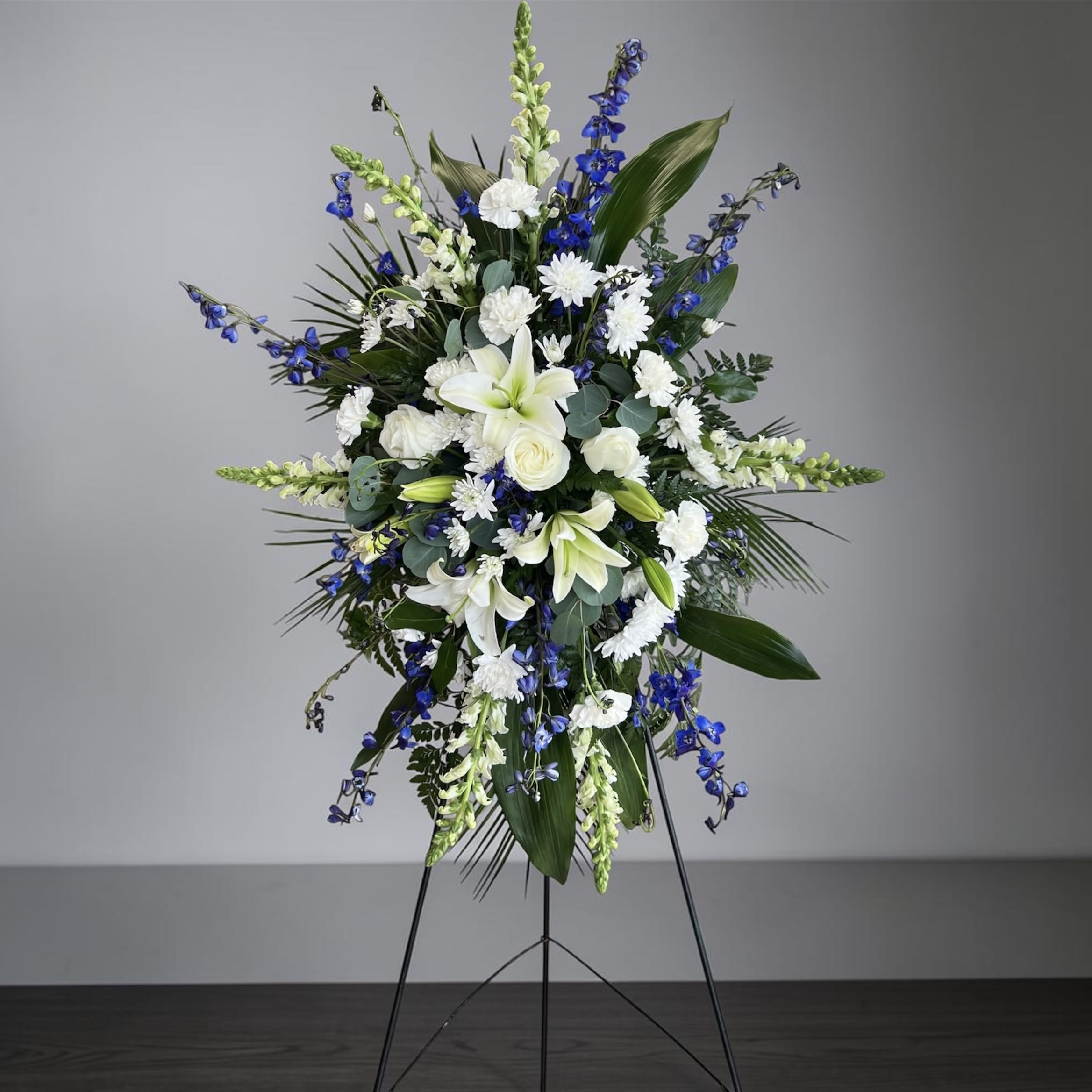 Peaceful Breeze Standing Spray  - A ceremonious display of white lilies, roses, carnations and chrysanthemums accented with vivid blue delphiniums and white snapdragon spikes-this standing spray is designed to convey calm and respect for a sympathy or memorial service. The vertical silhouette and restrained white, blue, and green palette give the recipient a feeling of quiet comfort and thoughtful presence, the kind of arrangement that quietly honors and remembers.