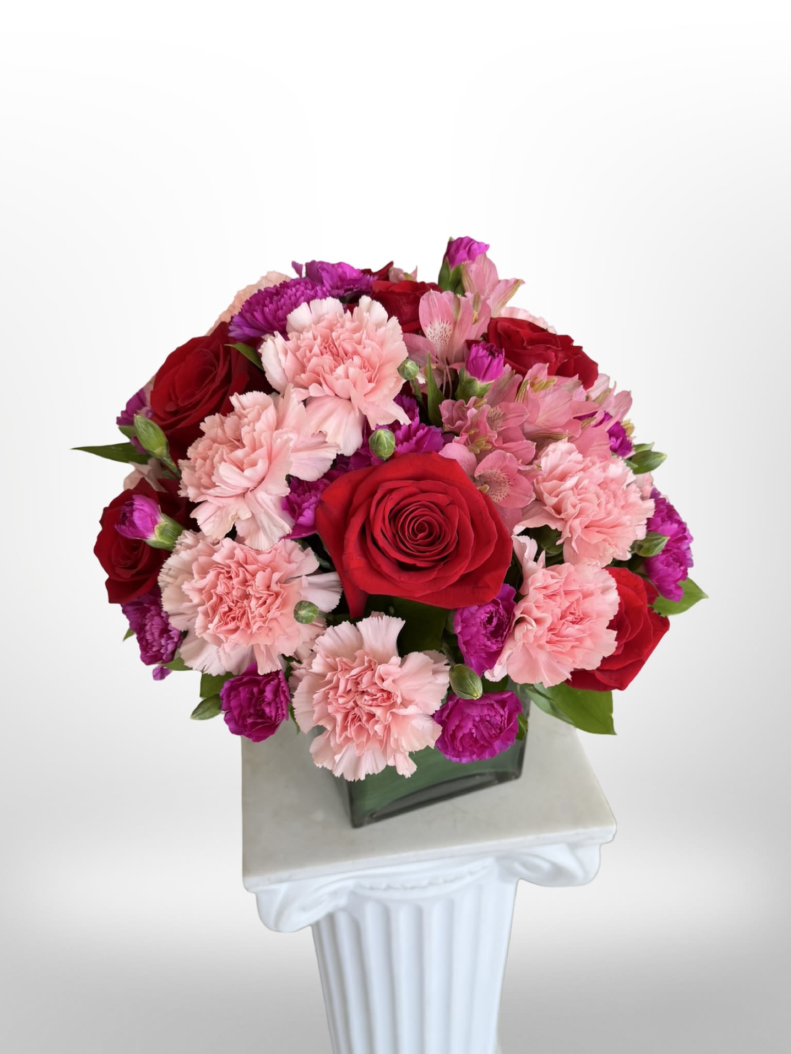 The Strawberry Cube  - A dense, rounded cluster of red roses and pale pink carnations gives this flower arrangement a punchy, romantic look - perfect as a birthday bouquet, anniversary roses gift, or a thoughtful everyday surprise. The compact silhouette and strong red-and-pink palette make the recipient feel celebrated and cherished, while bright magenta accents and touches of green add cheerful contrast that reads both joyful and slightly formal! 