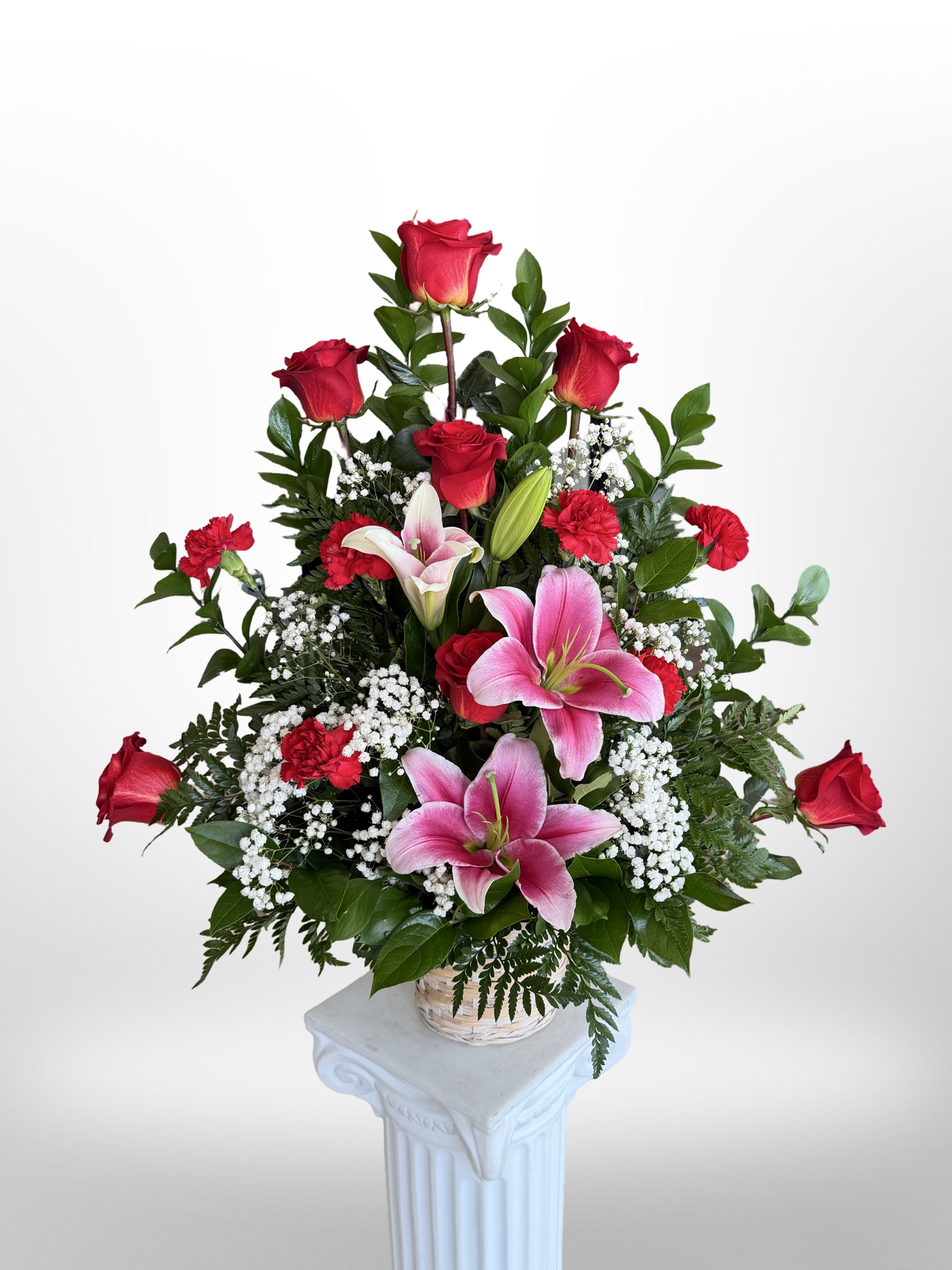 Ruby Basket  - A lush upright display of red roses and pink lilies, this basket arrangement reads as a classic romantic statement perfect for an anniversary bouquet, Valentine's Day gift, or a heartfelt sympathy spray. The combination of deep red and soft pink gives the arrangement immediate emotional clarity - it says love and comfort at once - while the airy white baby's breath and glossy greenery soften the silhouette for a tender finish! 