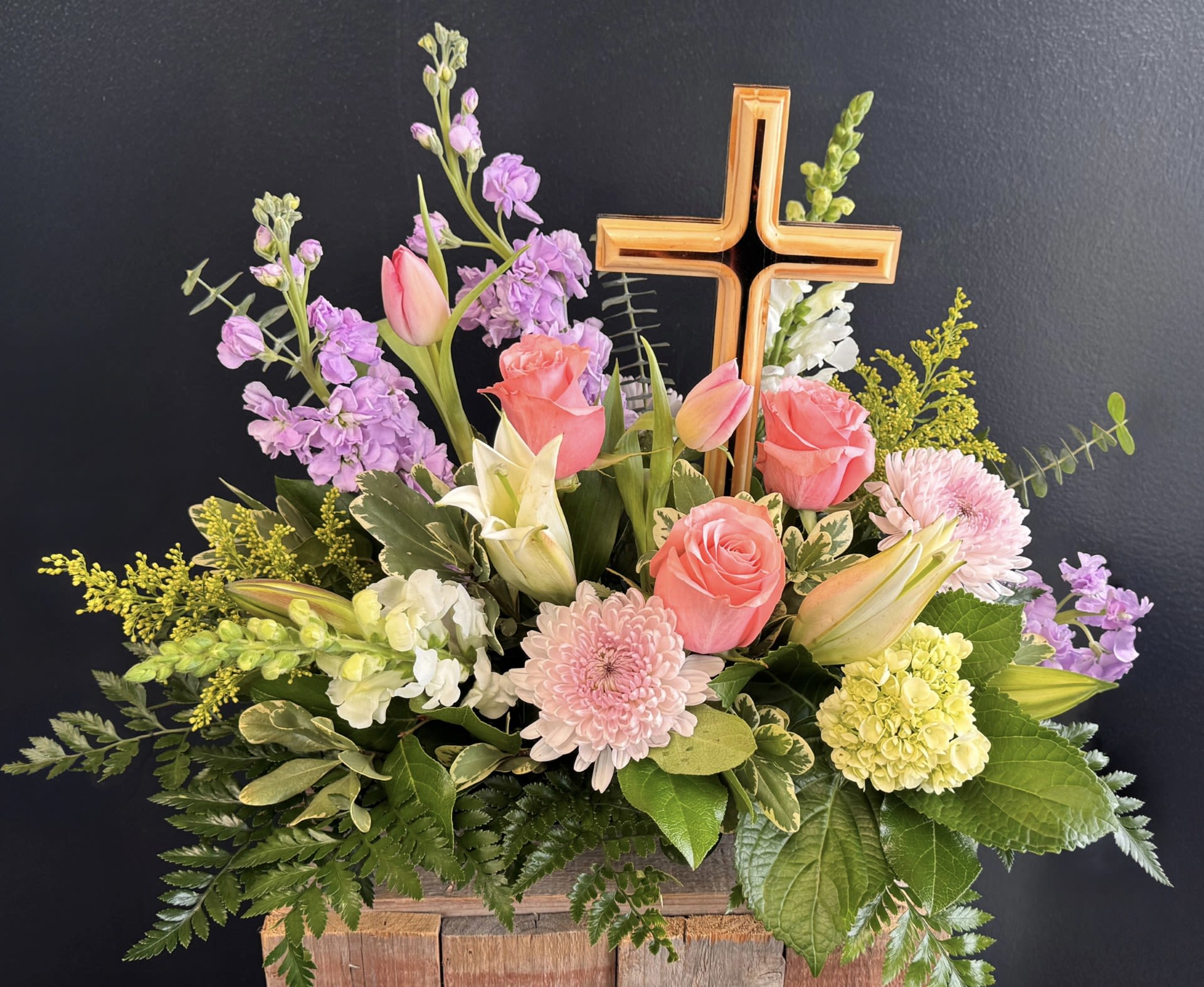 Forever in our Hearts  - A soft and elegant pastel floral arrangement designed to bring comfort and peace. Delicate blooms in shades of blush, lavender, cream, and soft peach surround a natural wooden cross, creating a beautiful expression of faith and remembrance. The combination of light, soothing colors and the rustic cross offers a heartfelt tribute, symbolizing hope, love, and eternal rest. Perfect for honoring a loved one with grace and serenity.