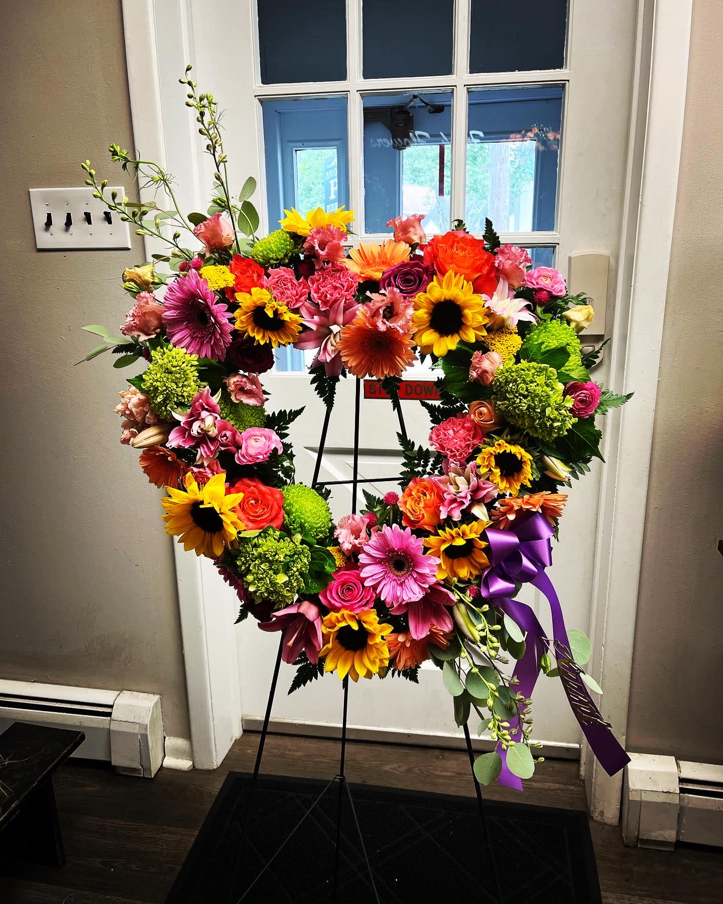 Heart-shaped floral wreath on a stand with bright mixed blooms and a purple ribbon
