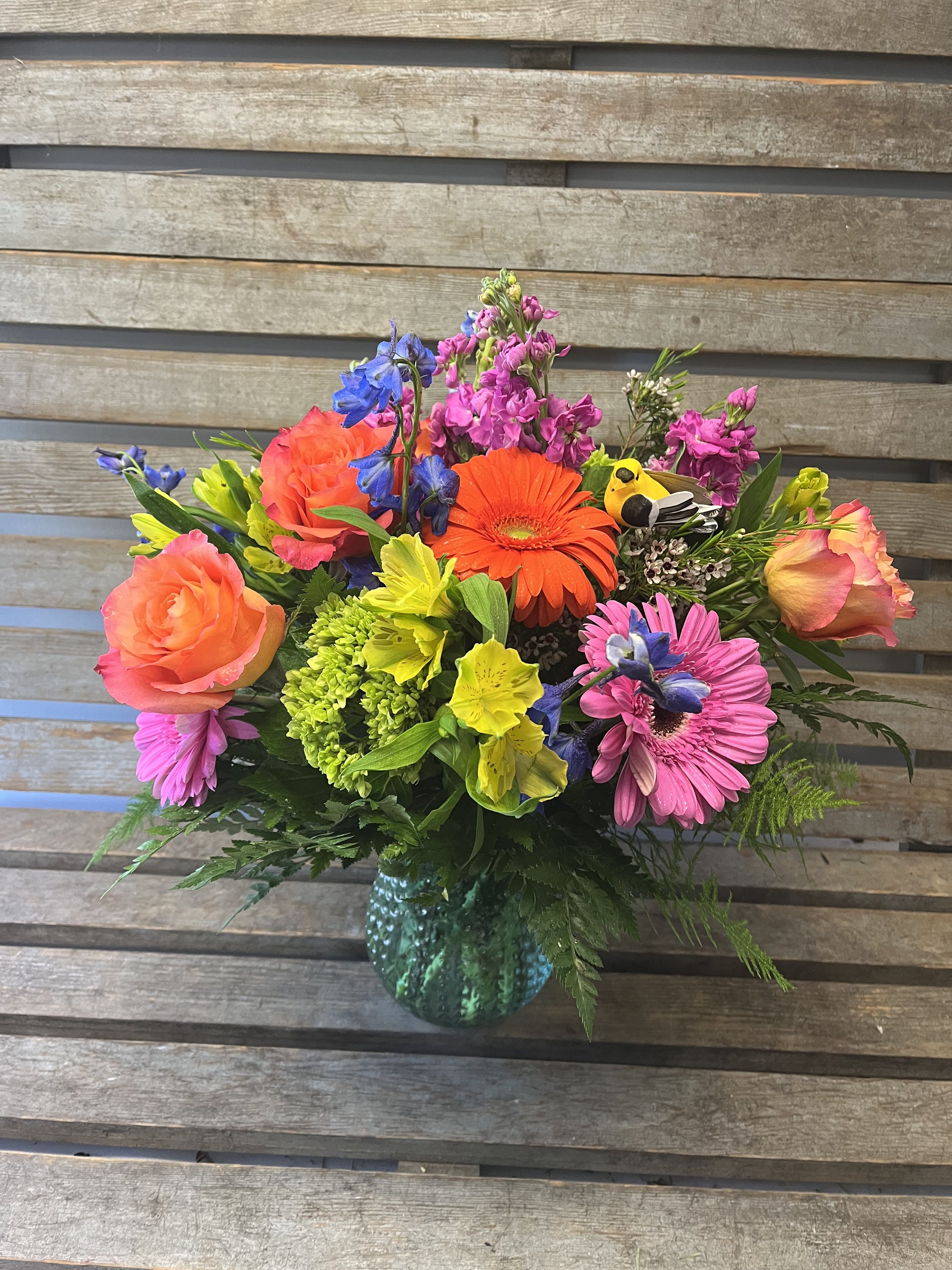 Bright  Designer's choice in ASST COLORED  glass vase ( VASE STYLE DIFFERS)  - Vivid gerbera daisies and roses in a teal glass vase create a joyful birthday bouquet that feels like a burst of sunshine - perfect for birthdays, congratulations, or an anniversary surprise. The mixed-color arrangement reads playful and uplifting, an ideal "just because" or get-well gift that makes the person you're thinking of feel genuinely seen and celebrated.  This designer's-choice arrangement combines large gerbera daisies with roses, alstroemeria, stock and other seasonal mixed blooms in a bright palette of yellow, blue, pink, orange and mixed tones. The flowers are gathered in a textured teal glass vase (vase style may vary) for a loose, garden-style silhouette that looks full and abundant on arrival. Seasonal accents such as pussy willow or a decorative butterfly may be included when available.  Whether you're sending love and romance, welcoming a new baby, saying thank you, or cheering on a graduate, this lively bouquet suits spring and summer occasions without feeling limited to a season. Sizes and exact flower varieties vary by availability; for the freshest presentation, the designer selects peak blooms at the time of delivery.
