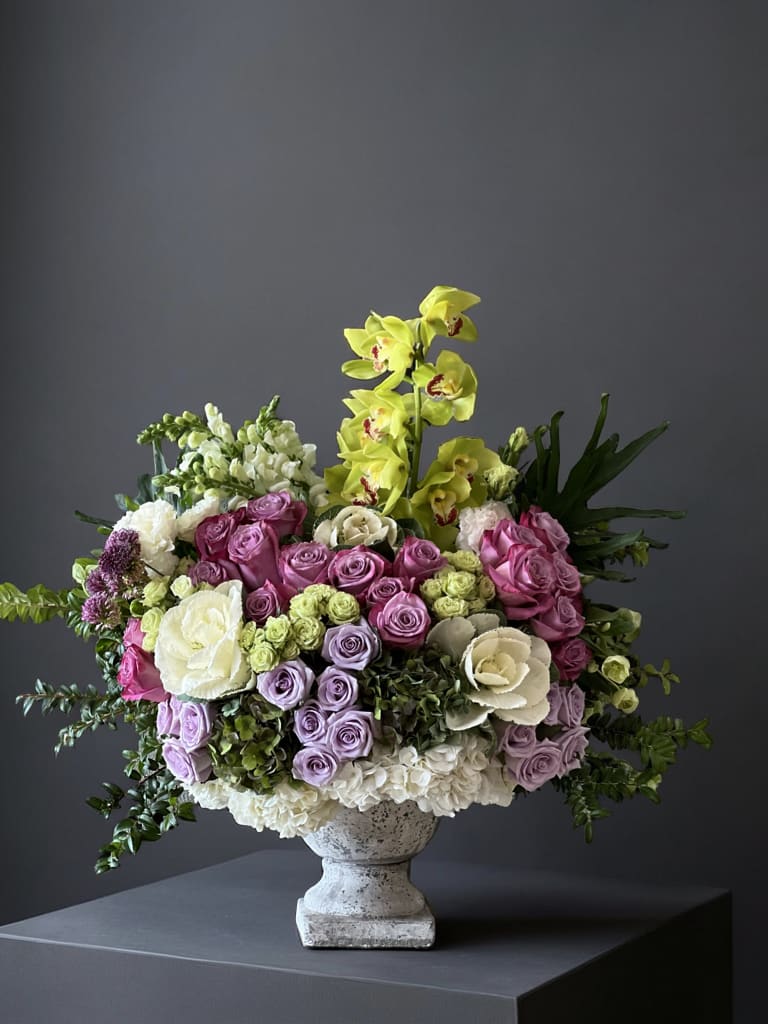 Garden Royale - An elegant garden-style arrangement featuring soft lavender and purple  roses, creamy hydrangea, delicate seasonal blooms, and striking green cymbidium orchids. Designed in a classic pedestal vase, this lush composition brings timeless beauty and refined charm to any space