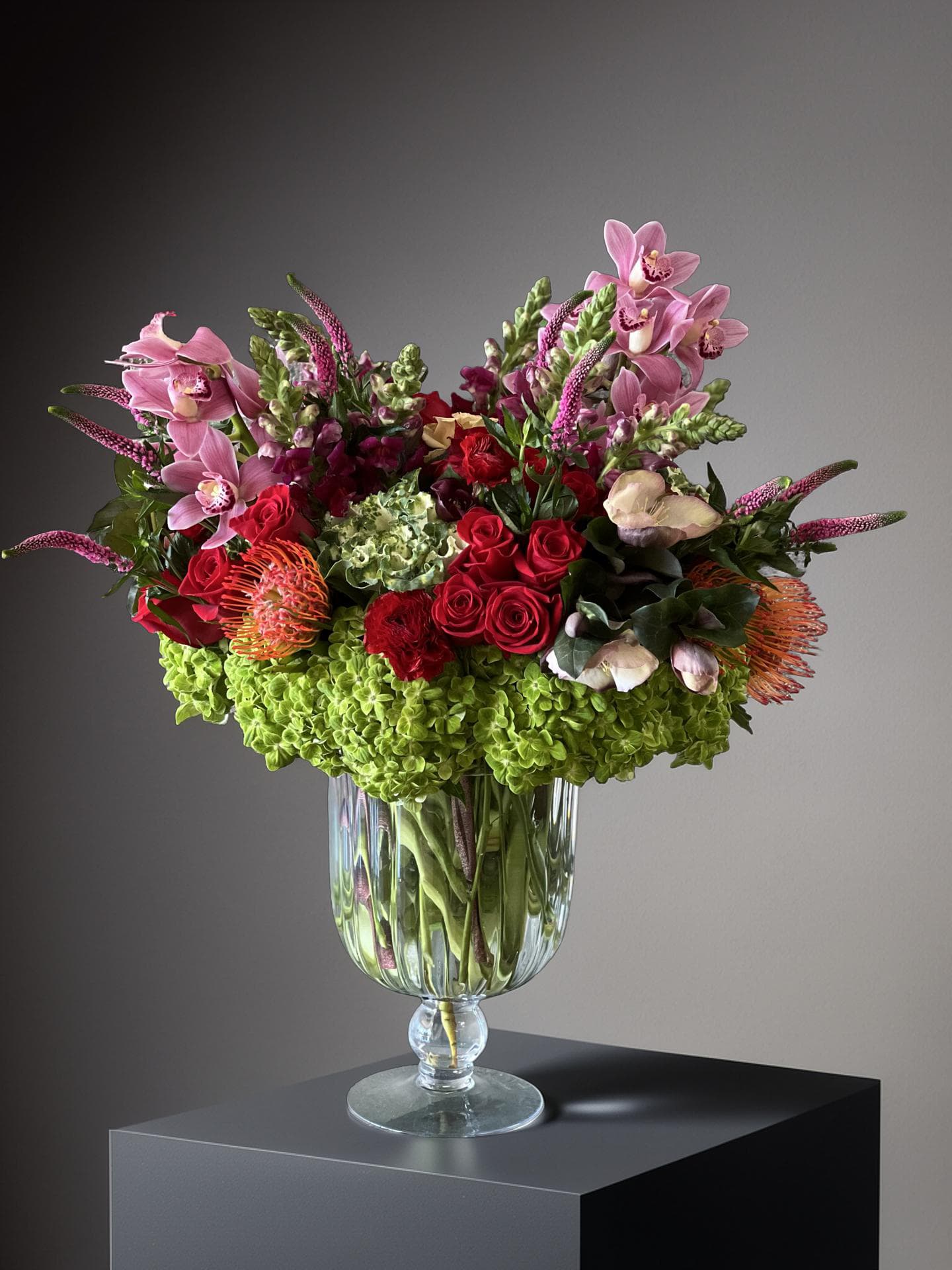 The Velvet Petal - The arrangement features a lush variety of blooms, including red roses, pink orchids, pincushion protea, green hydrangea, and pink veronica. The combination creates a romantic and bold statement, blending different textures and colors for an eye-catching display. This type of arrangement is ideal for sending as a memorable gift or brightening a special space.