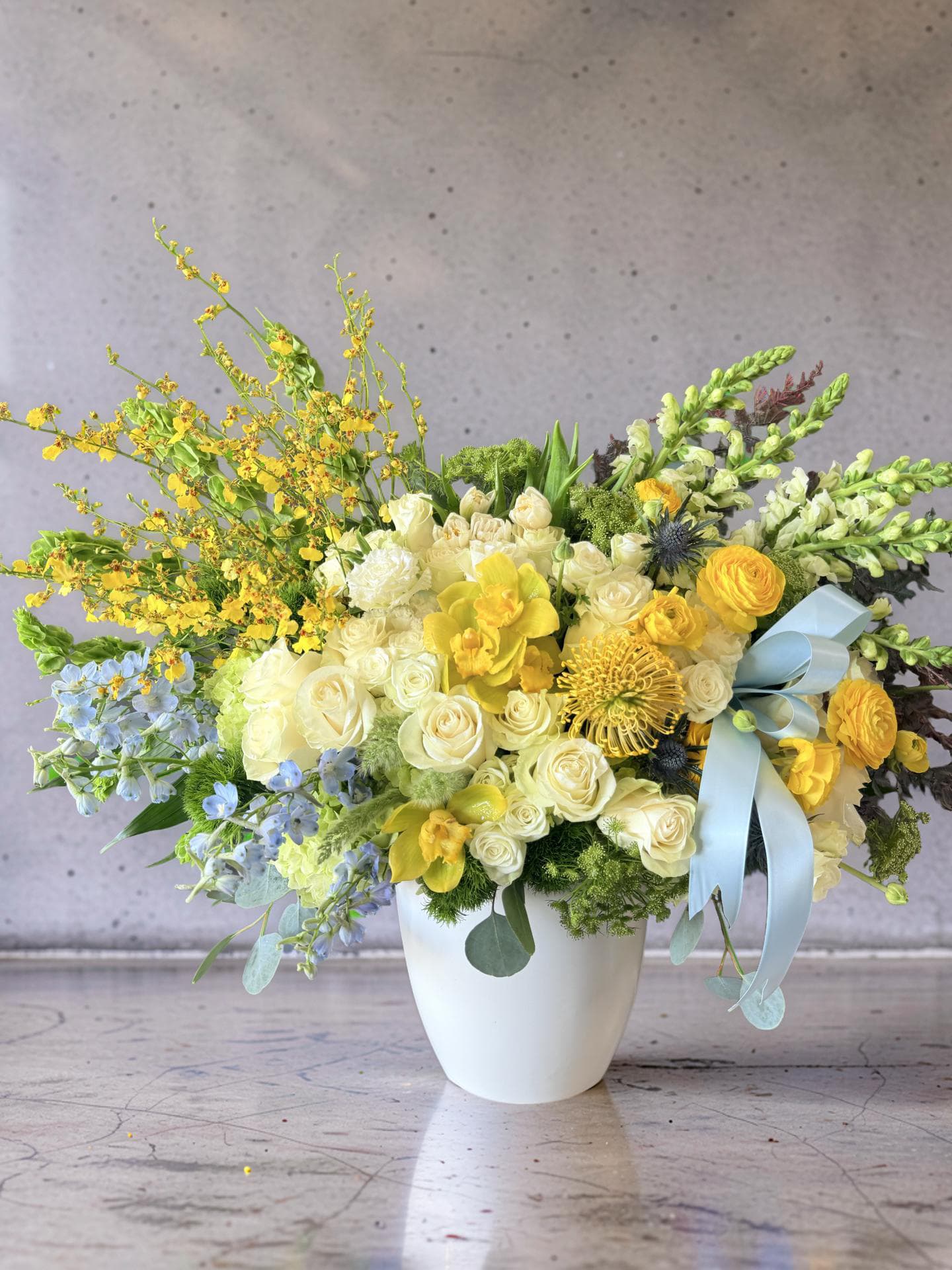 The Royal Oak - This stunning arrangement radiates happiness with a blend of sun-kissed yellow, crisp white, and soft blue colors, creating a fresh, garden-inspired look. It features a lush mix of roses, daffodils, delphinium, and other seasonal blooms, accented with a light blue ribbon and presented in a chic white vase. It is designed to impress with its volume and rich design.