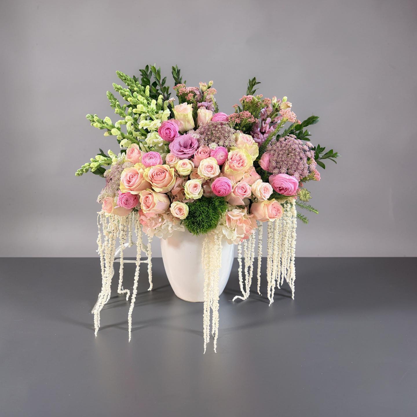 Blush Cascade Garden - A romantic and airy floral arrangement featuring soft blush and pastel tones, this design blends lush roses, ranunculus, and delicate seasonal blooms with fresh greenery. Cascading strands of amaranths create an elegant, flowing effect, adding movement and a touch of luxury. Designed in a modern white vessel, it’s perfect for weddings, celebrations, or refined home décor.