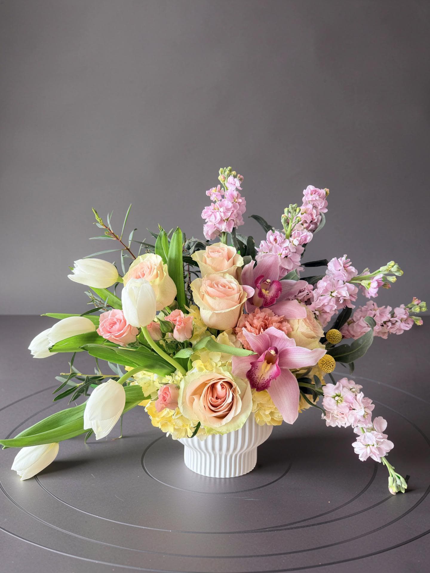 Blushing Love for Mother - Soft blush and peach or pink roses mingle with white tulips and pink orchids in a gentle pastel bouquet perfect for a Mother's Day bouquet or a birthday surprise. The arrangement's airy, garden-style silhouette and warm yellow hydrangea accents create a romantic, feminine feel that also works wonderfully for anniversaries, thinking-of-you moments, new baby welcomes, or a "just because" gift.  This flower arrangement features roses, tulips, orchids, stock, hydrangeas, and carnations arranged in a white ribbed ceramic vase. Pale stock and soft yellow hydrangea clusters add texture while several stems extend outward for a loose, graceful profile that reads fresh and relaxed. Sized to sit proudly on a dining table or bedside, it brings soothing pastels and a light, floral scent to brighten recovery rooms, graduation parties, housewarmings, or holiday gatherings.  Choose this arrangement to make the person you're thinking of feel truly seen-whether you're marking Mother's Day, saying thank you, sending get-well wishes, celebrating a retirement, or honoring a memory. Care is simple: keep water fresh, trim stems every few days, and display away from direct heat to prolong the life of the blooms.