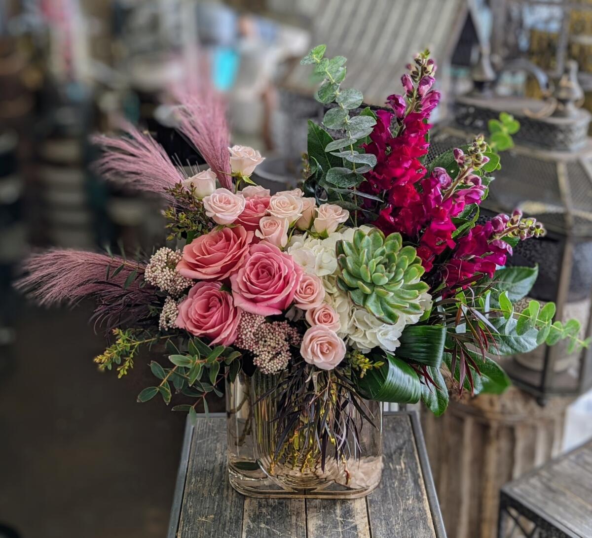 Eve - This floral arrangement is a striking fusion of elegance and modern flair, set in a sleek amber glass vase with a rounded rectangular form. It features a sophisticated blend of soft and bold elements, starting with lush pink roses and delicate blush spray roses as the centerpiece, softened further by white hydrangeas that add volume and texture.  Burgundy snapdragons draw the eye upward with their height and rich color, adding dramatic vertical interest. A large succulent nestled among the blooms adds a unique, contemporary twist and a textural contrast.  Accents of soft pink pampas grass, wispy seeded eucalyptus, burgundy foliage, and structured greenery round out the design, giving it movement, structure, and depth.  This arrangement is perfect for someone who loves a mix of classic romance with a trendy, natural twist—ideal for birthdays, celebrations, or stylish décor.  Creative and inspired arrangements handcrafted in Las Vegas. Same day delivery available. 