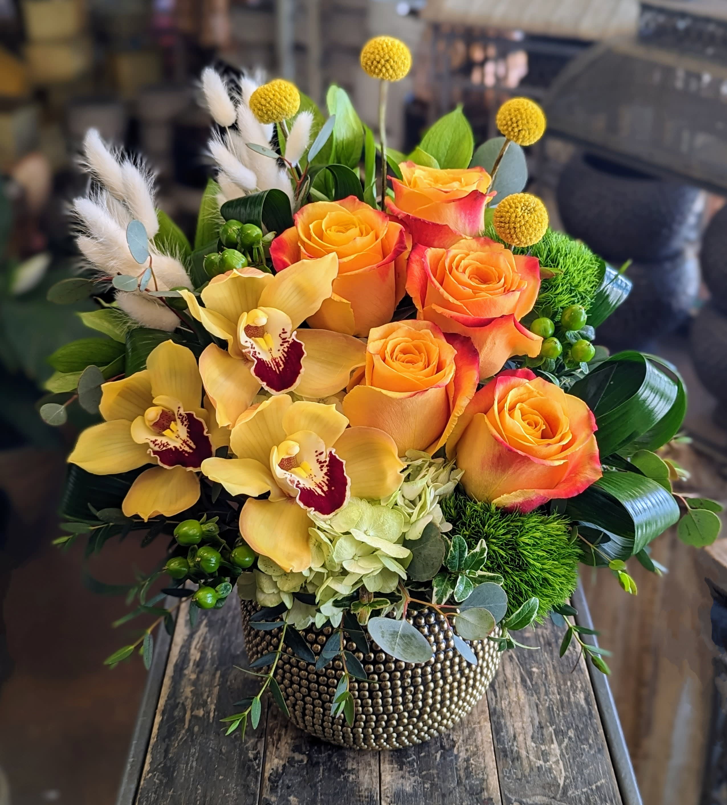 Tangerine - Tangerine Bold, bright, and full of joy, Tangerine is a modern floral arrangement from Windmill Floral Expressions in Las Vegas, handcrafted to spark warmth and delight. Designed in a textured gold vessel, this radiant bouquet blends tropical flair with contemporary elegance, making it a standout for any celebration.  At its heart, orange and yellow bi-color roses glow with citrusy brilliance, while exotic yellow cymbidium orchids add a touch of lush sophistication with their deep burgundy centers. Cheerful yellow billy balls rise playfully above the design, contrasted by soft white bunny tails that bring a whimsical, airy texture.  Grounded by fresh green moss, hypericum berries, and pale green hydrangea, Tangerine is finished with elegant loops of foliage and sprigs of eucalyptus for a polished, full look. Bursting with personality and golden-hour charm, this arrangement makes the perfect gift or statement piece.  Las Vegas Luxury Florist – Sophisticated Floral Designs for the Most Elegant Moments. Same-day delivery available.