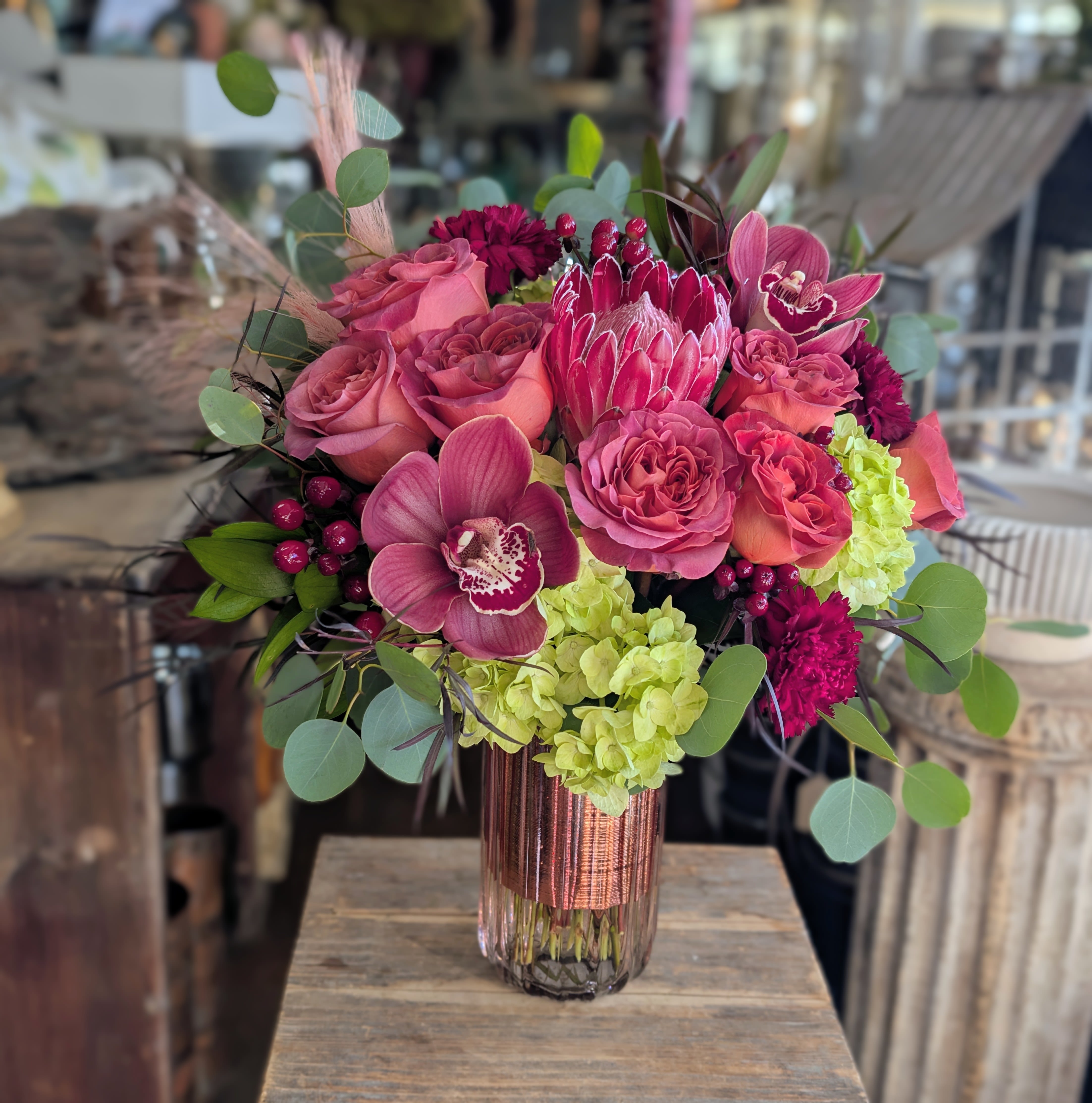 Isadora - Isadora is a free-spirited, bohemian floral arrangement designed to feel artful, romantic, and effortlessly collected. This lush composition features layered garden roses, cymbidium orchids, striking protea, soft hydrangea, seasonal berries, and textural greenery—each bloom thoughtfully placed to create natural movement and depth. Warm berry and rose tones mingle with fresh green accents, all gathered into a glowing blush vessel that adds richness and modern warmth. The result is an elevated yet organic design that feels both timeless and expressive, perfect for celebrations, anniversaries, heartfelt gestures, or moments that call for something truly special. Hand-crafted in our Las Vegas studio using premium flowers and designer techniques, Isadora reflects Windmill Floral Expressions’ signature blend of modern luxury and bohemian soul. Custom color palettes and flower substitutions are always available to suit your vision. Available for flower delivery throughout Las Vegas and Henderson. Same-day delivery available.