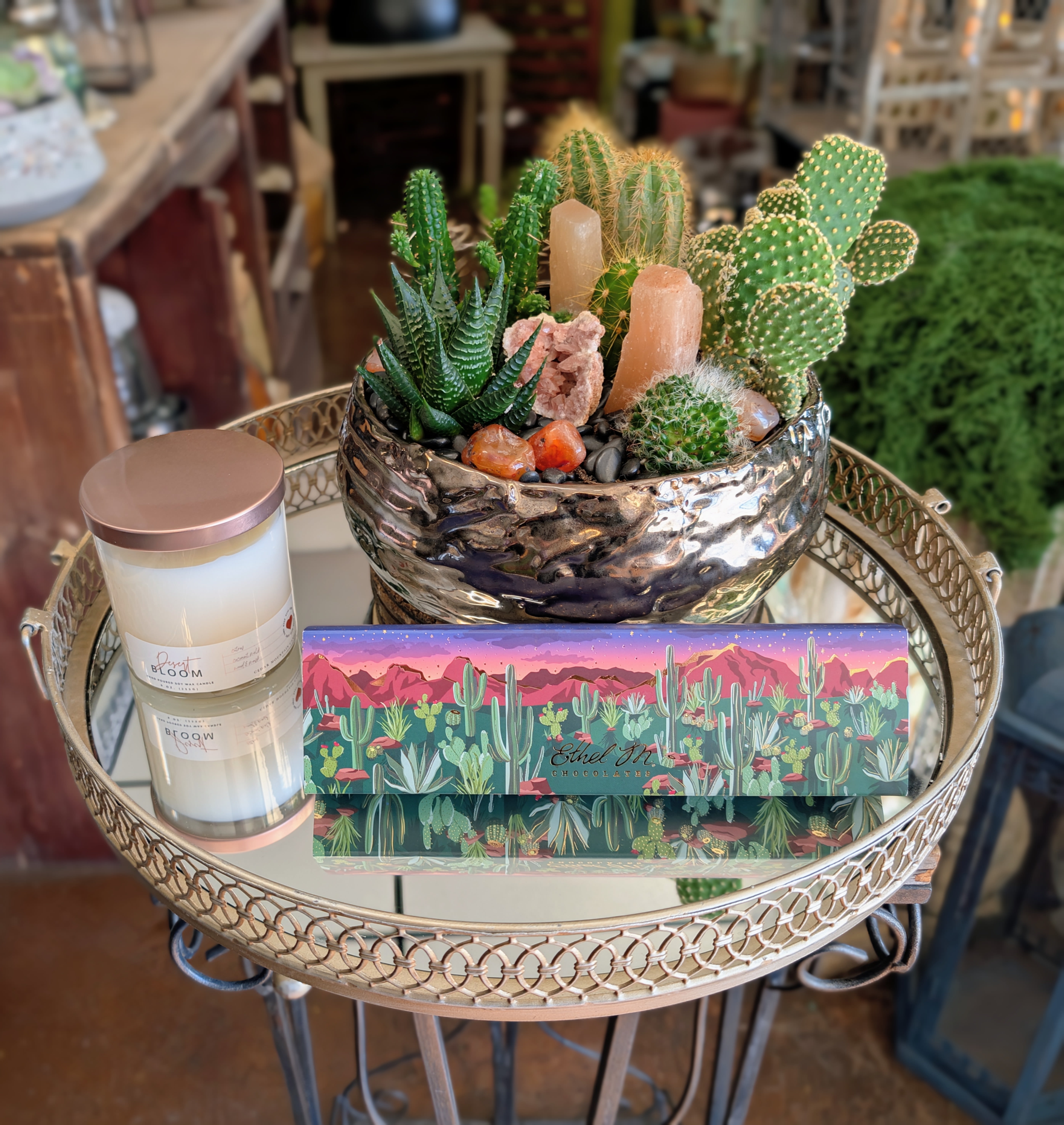 Sonoran Luxe Gift Cactus Garden- With A Desert Bloom Candle And Ethel M 5 Piece Chocolates - Bring the spirit of the Mojave Desert to life with our Sonoran Luxe Gift Cactus Garden, an elevated gift featuring a collection of living cacti and succulents nestled among natural crystals and sculptural selenite in a modern hammered metallic ceramic vessel.  Thoughtfully paired with a luxurious Desert Bloom candle by Cedar Mountain Candle and handcrafted 5 piece gourmet chocolates from Ethel M Chocolates, this gift celebrates the beauty, warmth, and artistry of the Southwest. It offers a memorable blend of living botanicals, relaxing fragrance, and iconic Las Vegas indulgence.  Perfect for birthdays, housewarmings, client appreciation, thank-you gifts, retirement celebrations, or anyone who appreciates distinctive desert-inspired design, this arrangement is both sophisticated and long-lasting.  Designed exclusively by Windmill Floral Expressions, each garden is individually created using premium cacti, succulents, natural crystals, and carefully selected decorative elements. Because every garden is handcrafted, plant varieties and crystal accents may vary slightly, making every piece truly one of a kind.  A beautiful luxury gift that captures the timeless elegance of the desert while bringing lasting beauty to any home or office.
