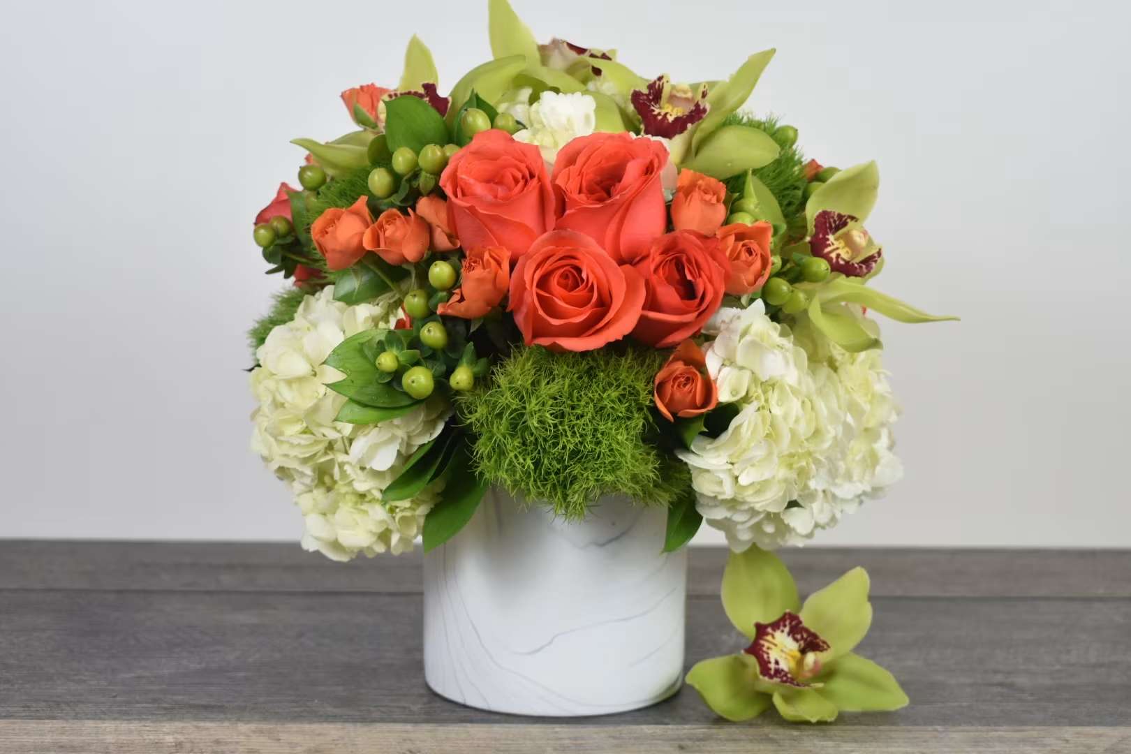 Low arrangement of orange roses, green orchids, and white hydrangeas in a white vase with one orchid bloom beside it.