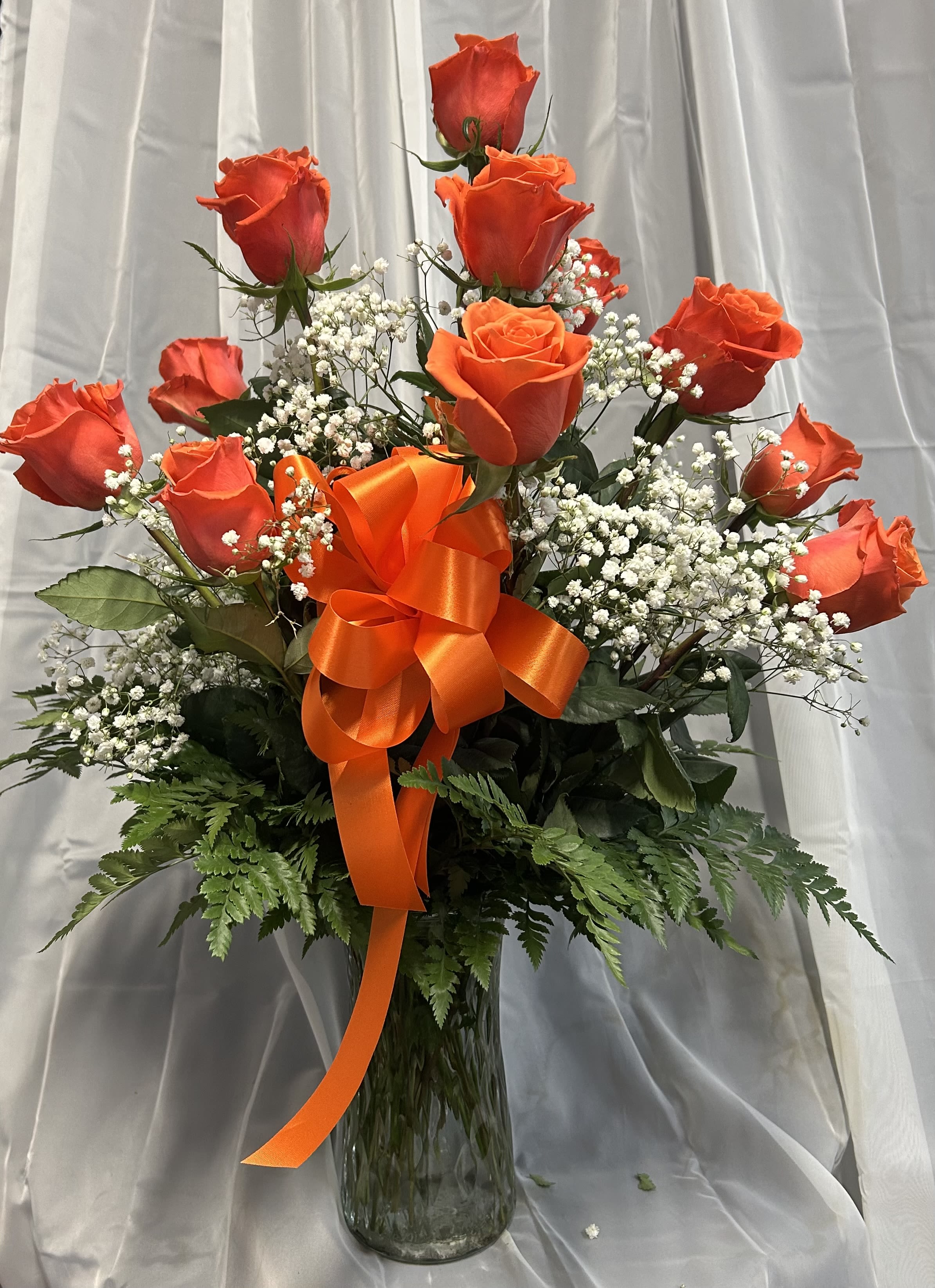 Orange roses in a glass vase with a large orange ribbon and white filler flowers.