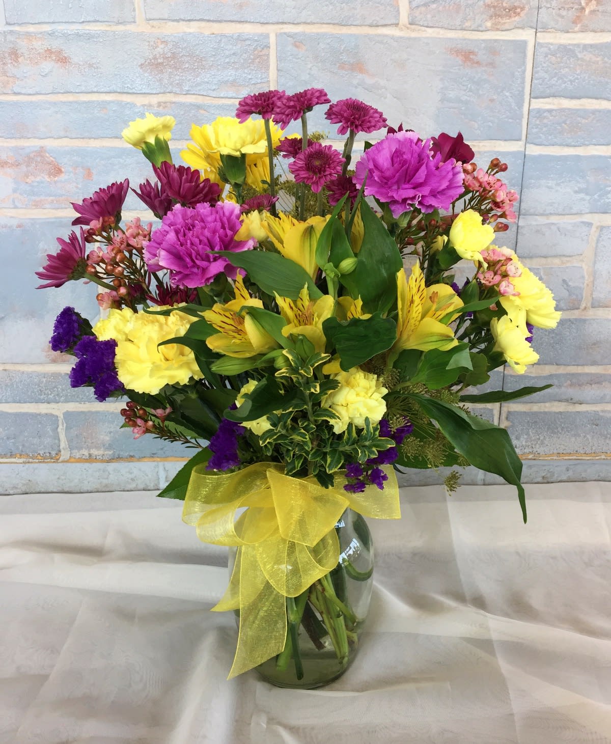 Mixed bouquet of yellow, pink, and purple flowers in a glass vase with a yellow ribbon