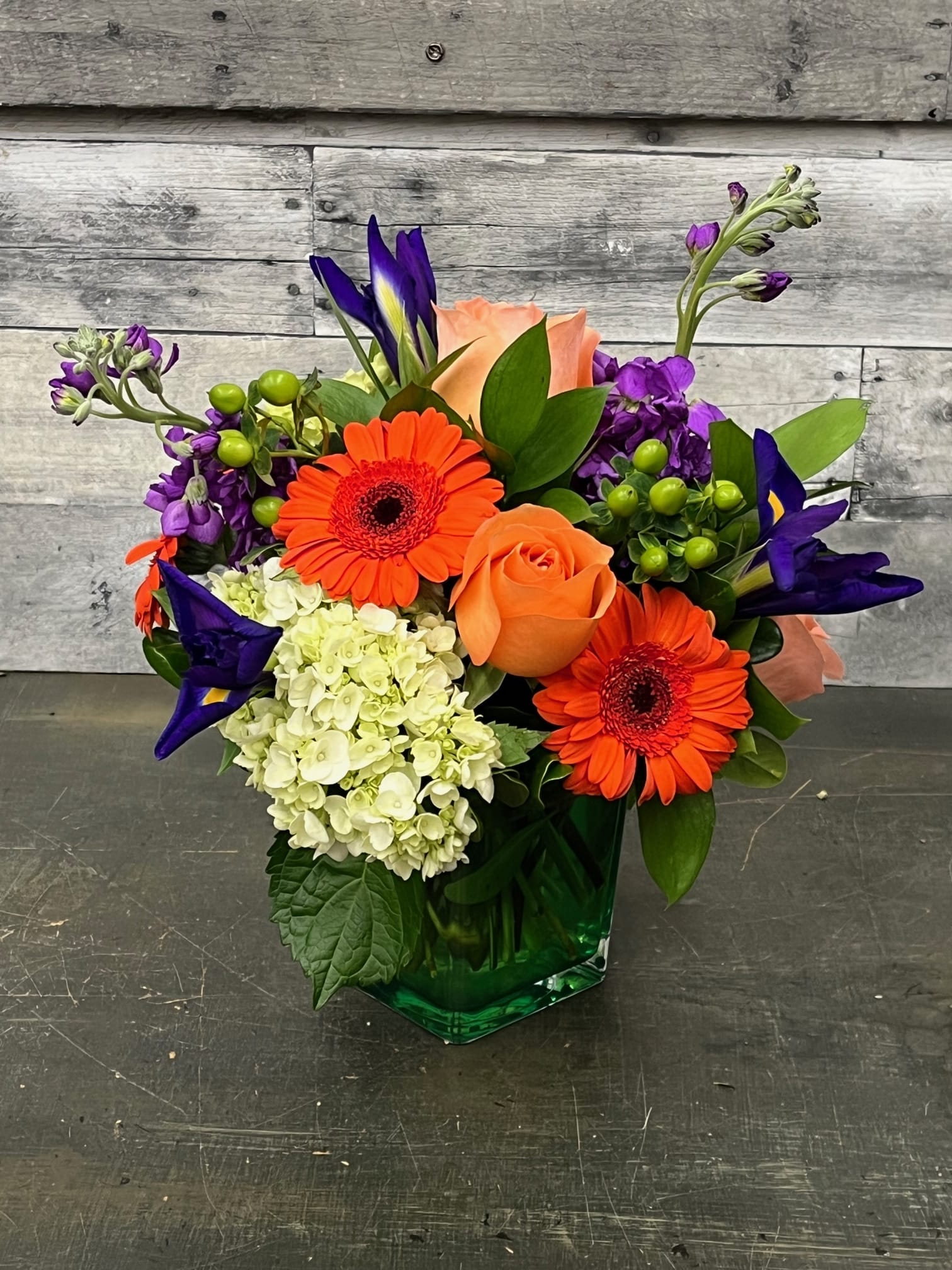 Bright mixed bouquet with orange gerberas, peach roses, and purple blooms in a green vase.