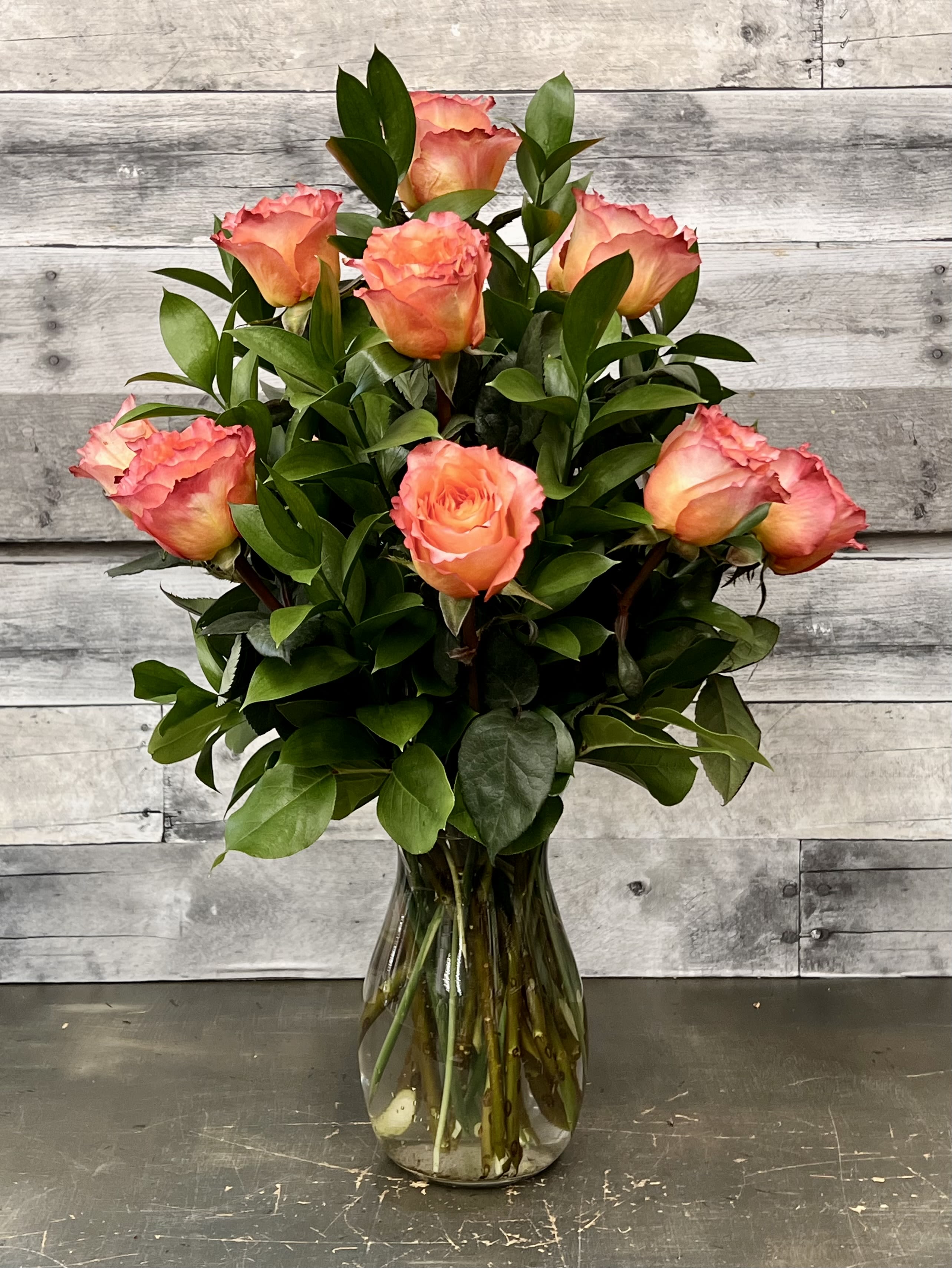 Classic Orange Roses - A radiant cluster of pink-orange roses with ruffled outer petals and warm peach centers creates an immediate sense of warmth and romance - perfect as a birthday bouquet, anniversary roses, or a thoughtful thank-you arrangement. The rounded, upright shape reads classic and polished, so the person you're gifting will feel genuinely celebrated the moment these arrive.  Arranged in a clear glass vase, the roses are cushioned by abundant dark green foliage that amplifies the color contrast and gives the bouquet a fresh, everyday elegance. The compact, approachable size works well on a dining table, desk, or bedside table; simple care - fresh water in the vase and trimming stems every few days - will keep the blooms looking their best while they brighten someone's day.
