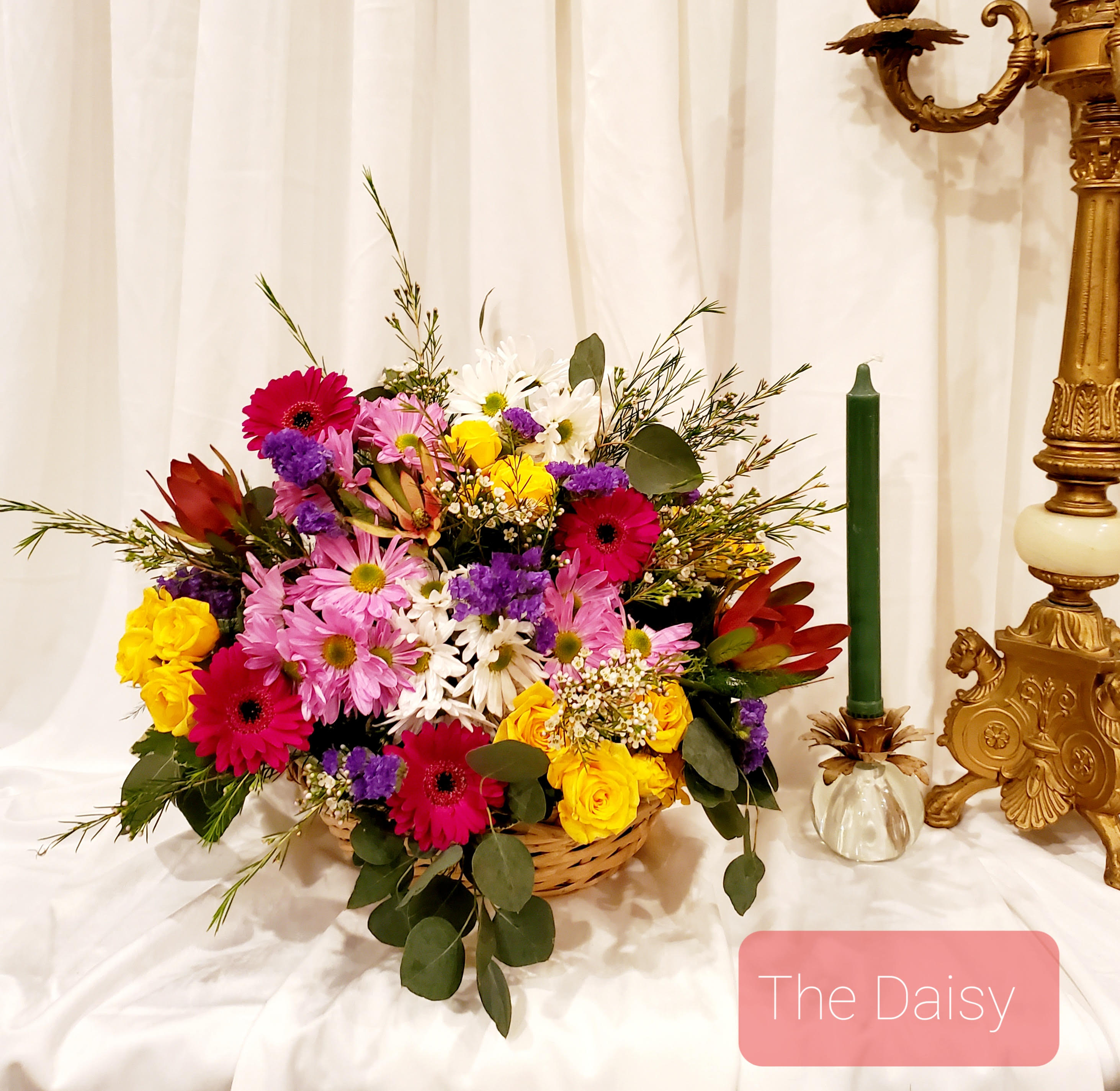 Basket arrangement of mixed flowers beside a green candle