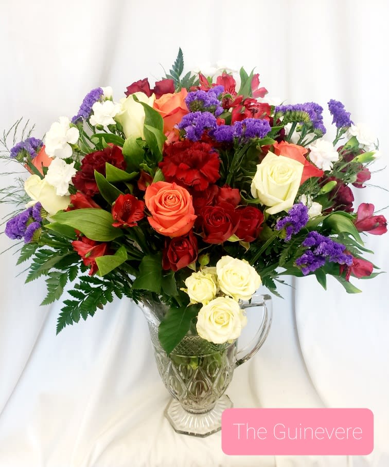 Mixed bouquet of roses, carnations, and purple filler flowers in a glass pitcher vase