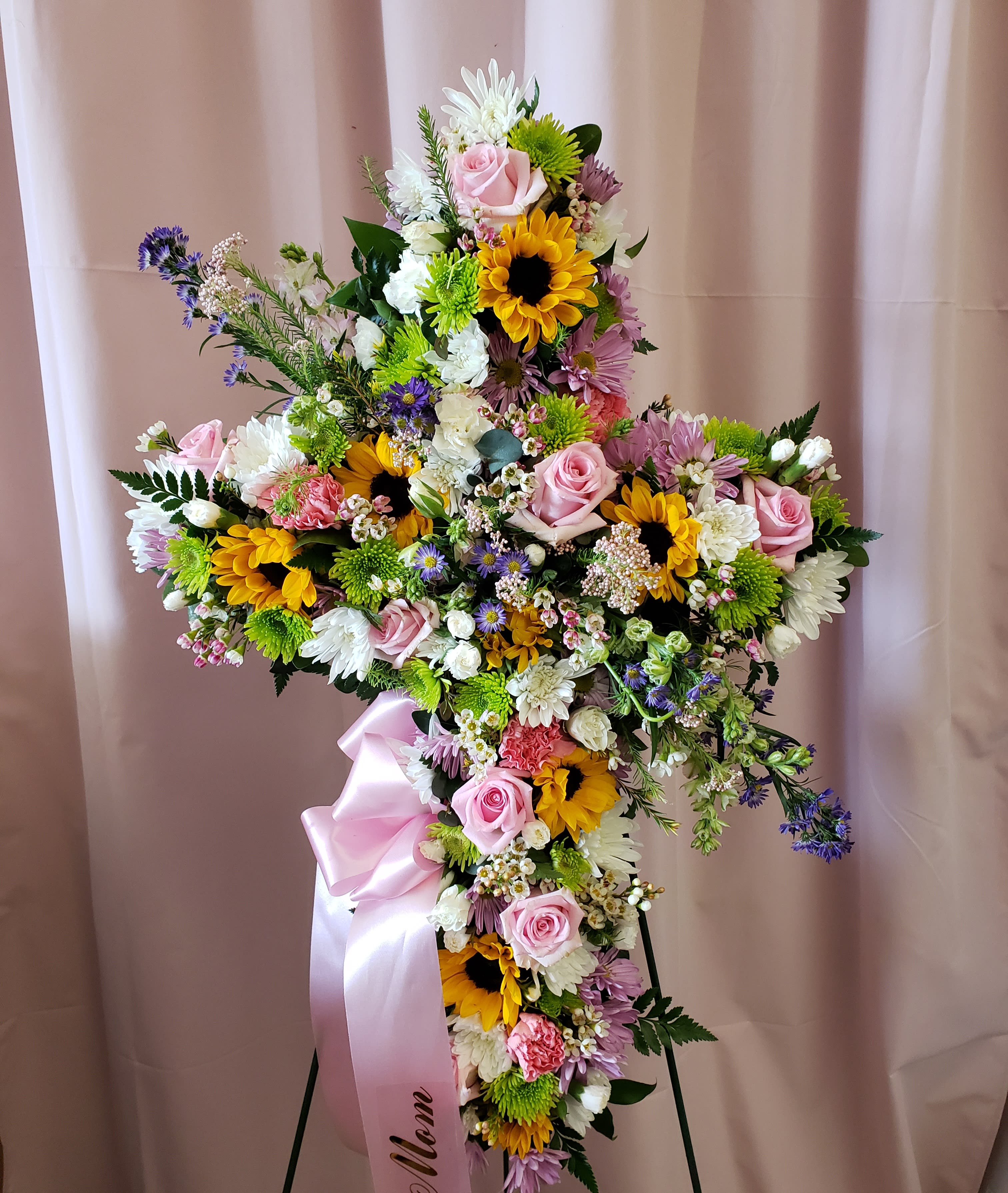 Large floral cross with pink roses, sunflowers, and white daisies on a stand