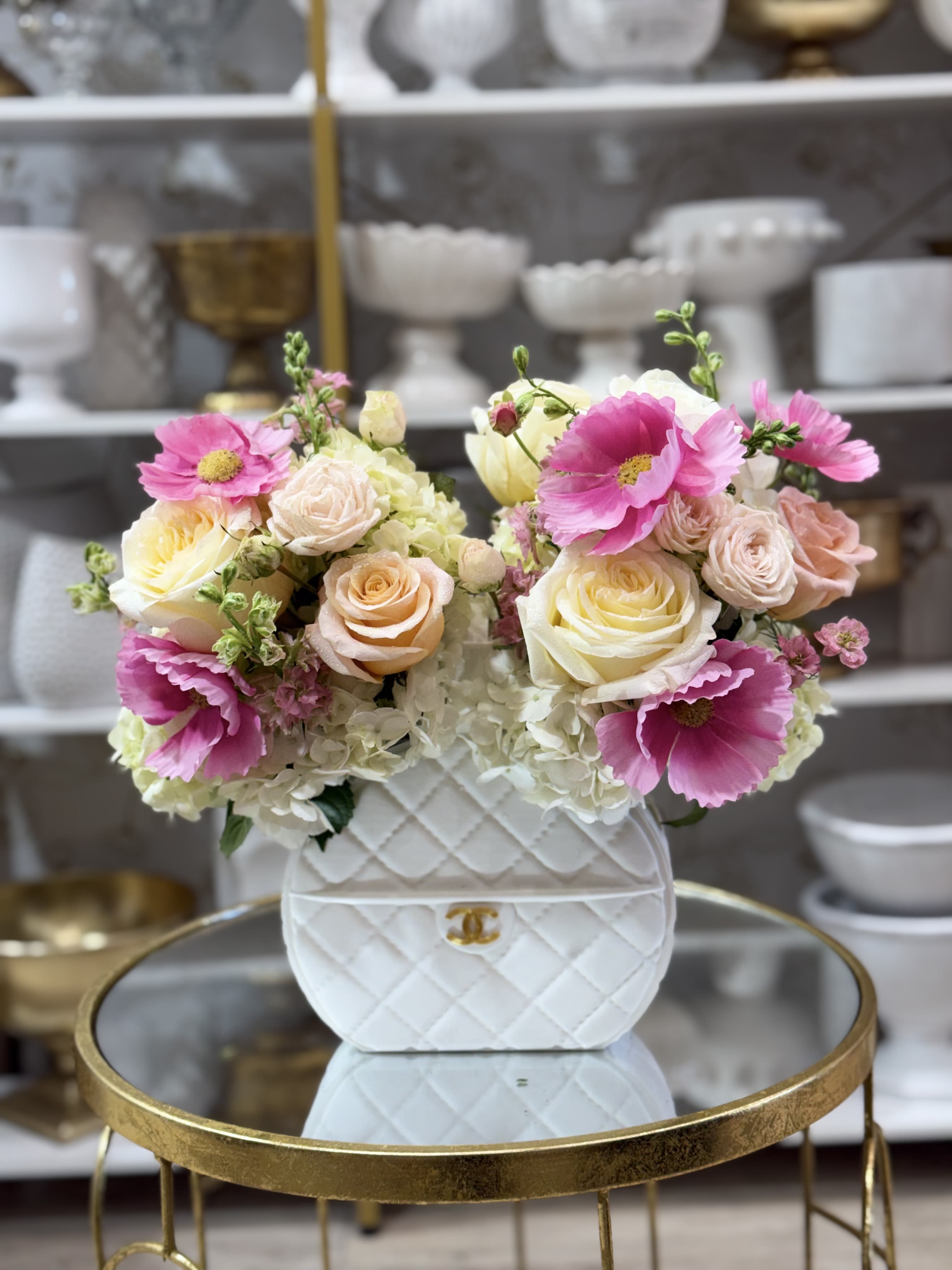 Handbag-shaped white ceramic vase filled with pink anemones, pastel roses, and white hydrangeas on a gold table