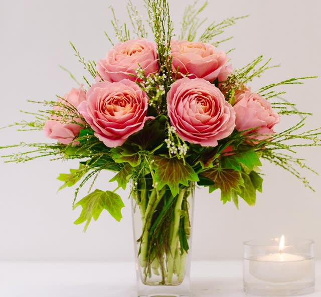 Pink roses arranged in a clear glass vase with greenery and a lit candle nearby