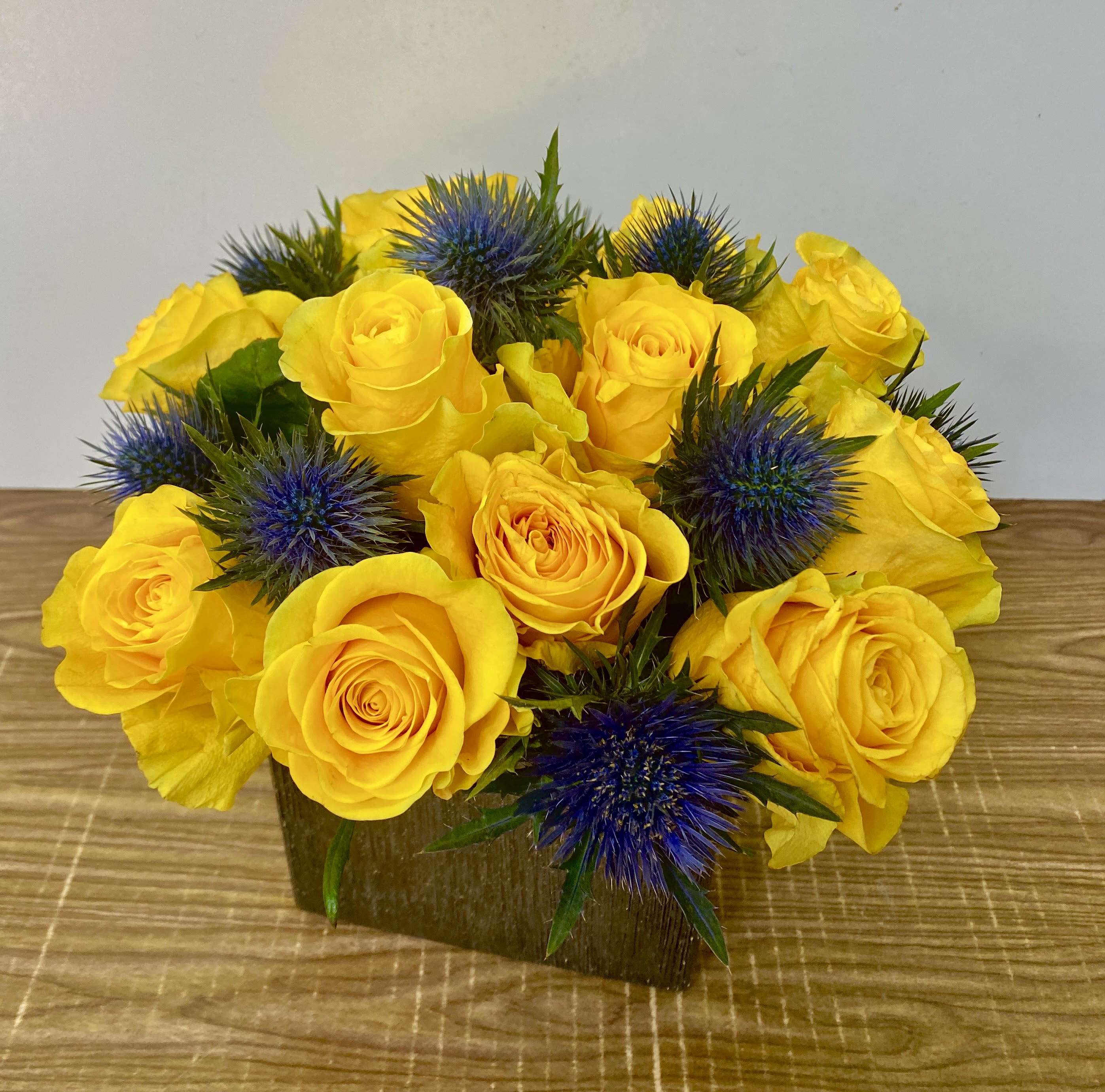 Yellow roses with blue thistle in a low square vase