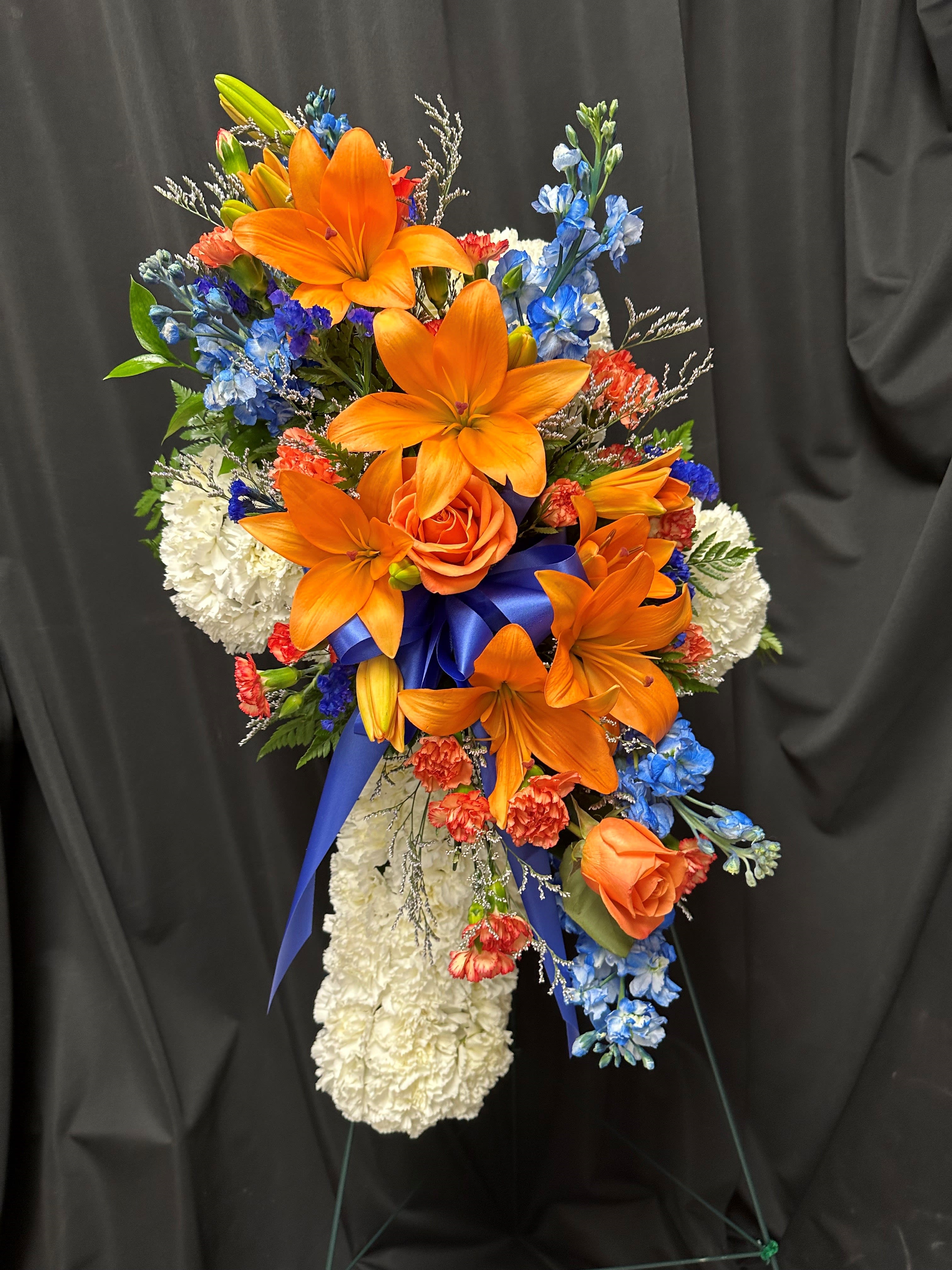 Tall cross-shaped spray of white carnations with orange lilies, roses, and blue flowers on a stand