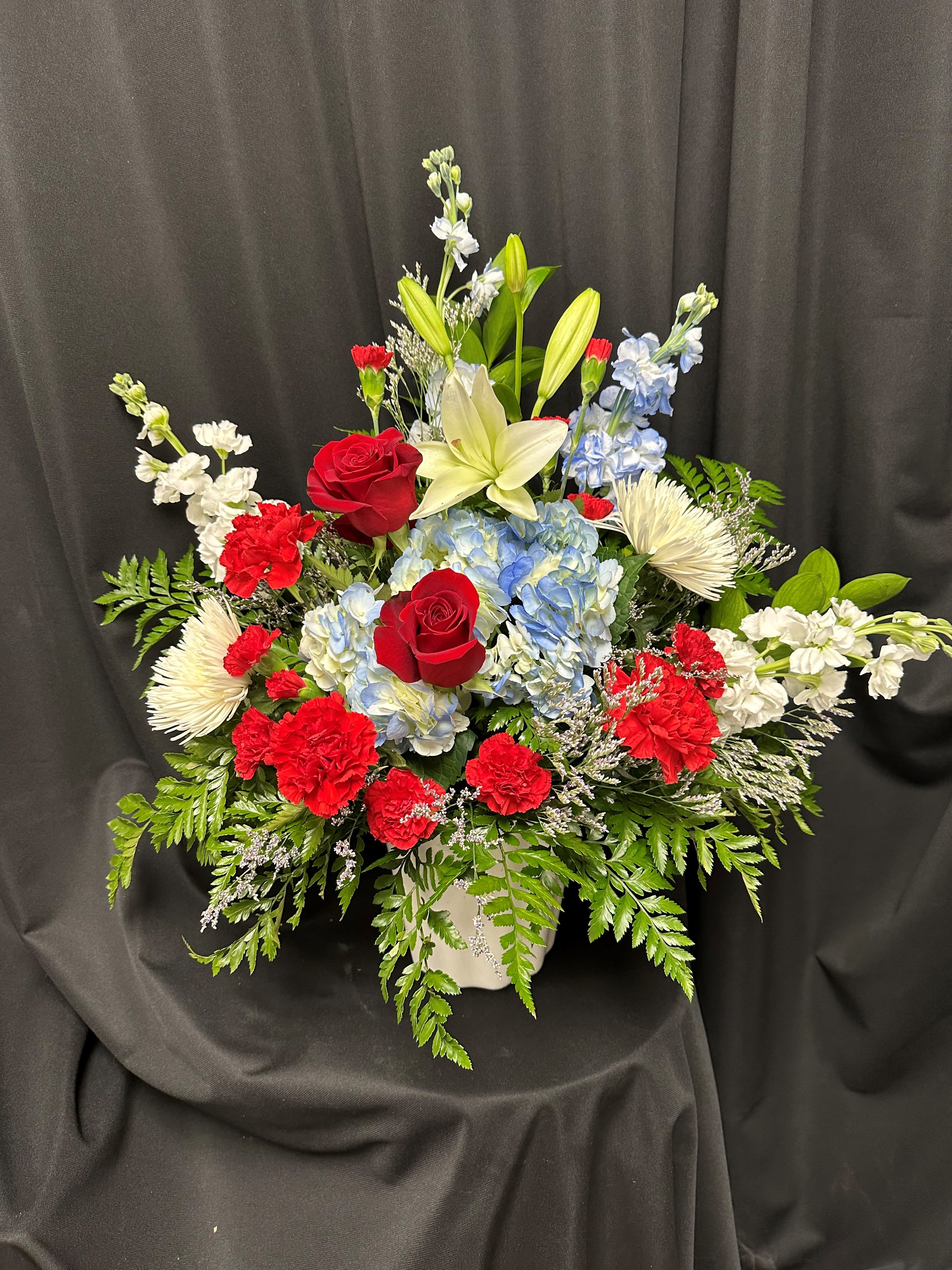 Honored Service - Pay tribute to the dedication and sacrifice of the service member in your life with this patriotic basket of fresh red, white, and blue blooms. Featuring the regal presence of blue hydrangeas, the passion of red roses, the purity of white mums and lilies, and the resilience of red carnations and white stock, each bloom honors their commitment with grace and gratitude. This heartfelt arrangement serves as a meaningful token of appreciation for their service and sacrifice, expressing admiration and respect in a beautifully symbolic way.