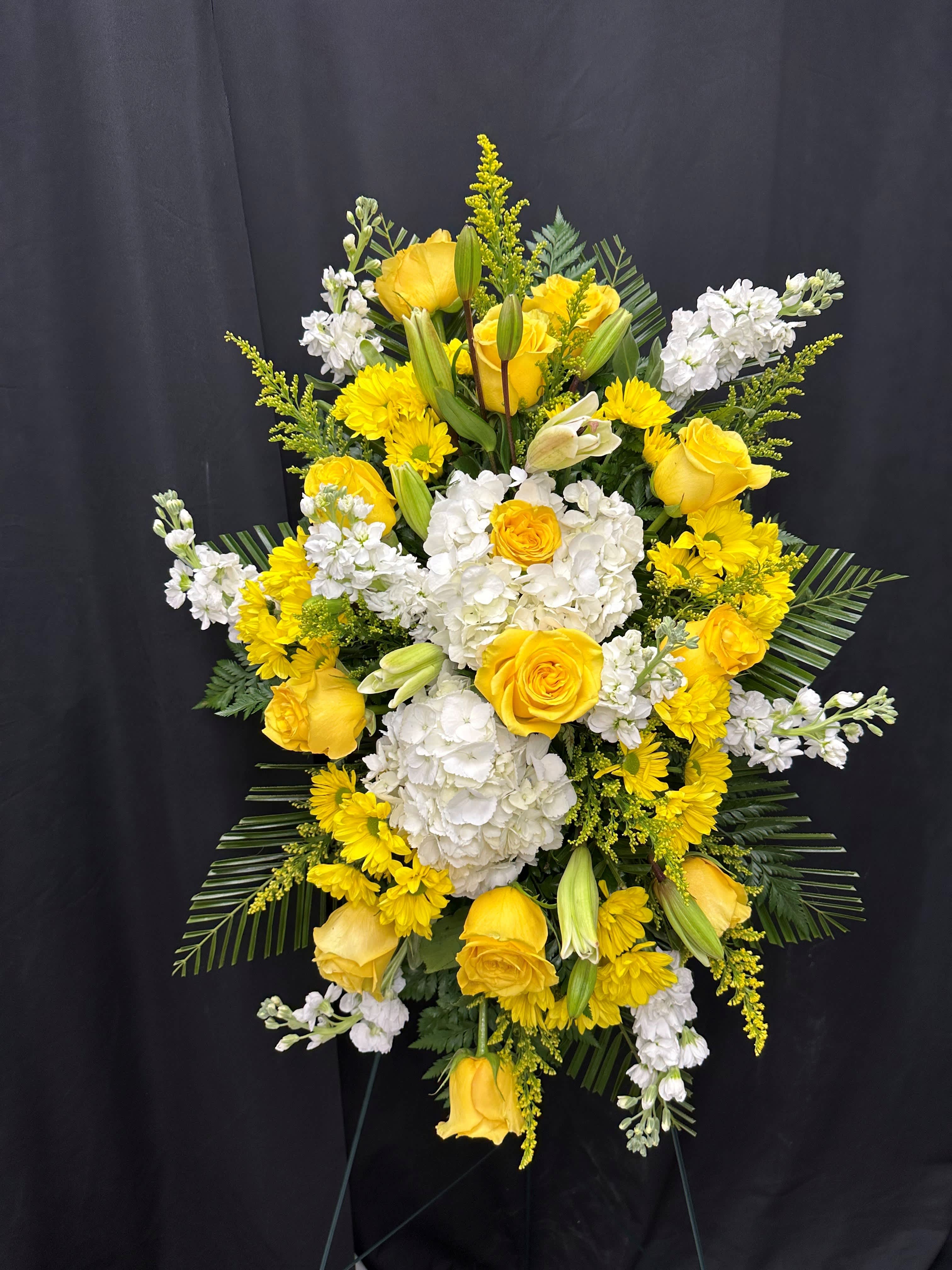 Tall standing spray of yellow roses, white hydrangeas, and yellow daisies on a green easel.