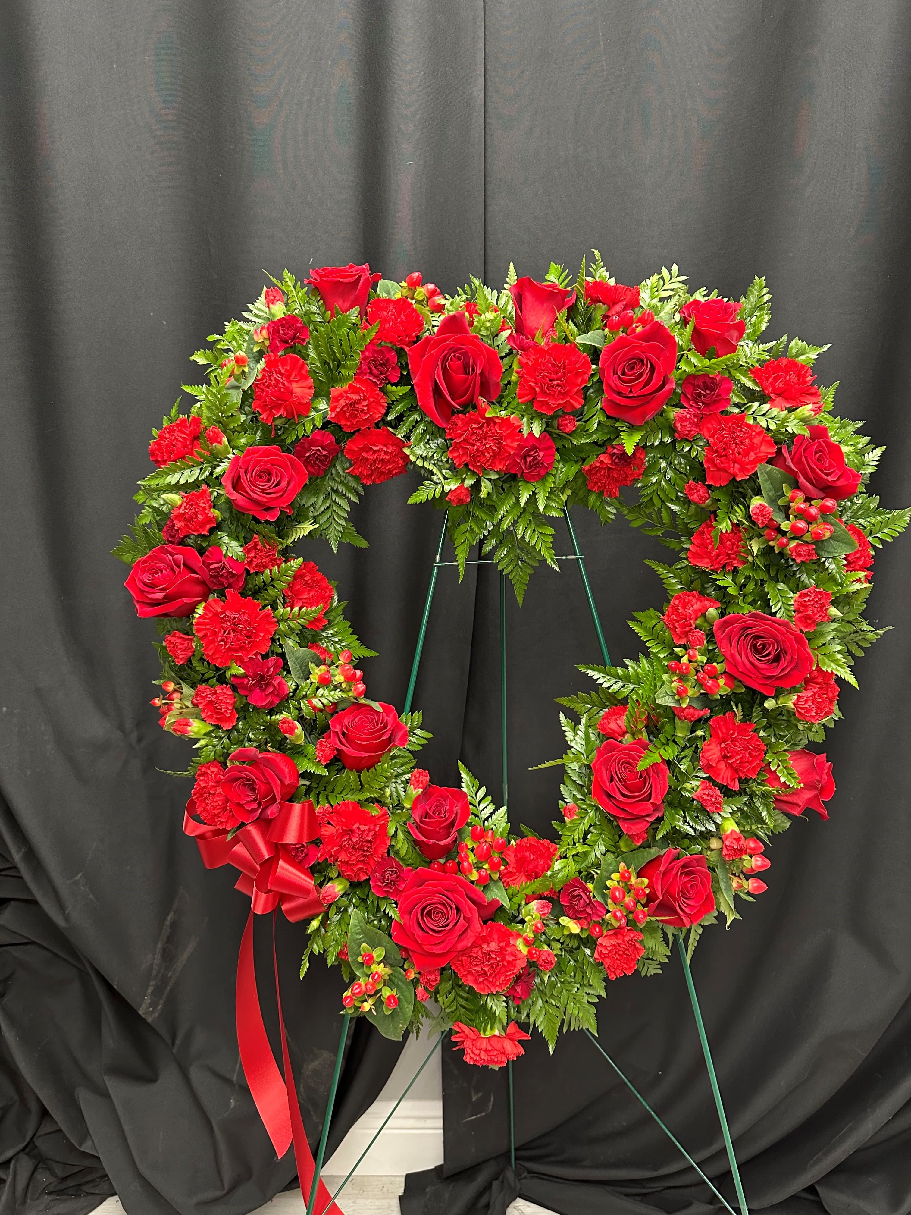 Heart-shaped red rose and carnation wreath on a stand with a red ribbon