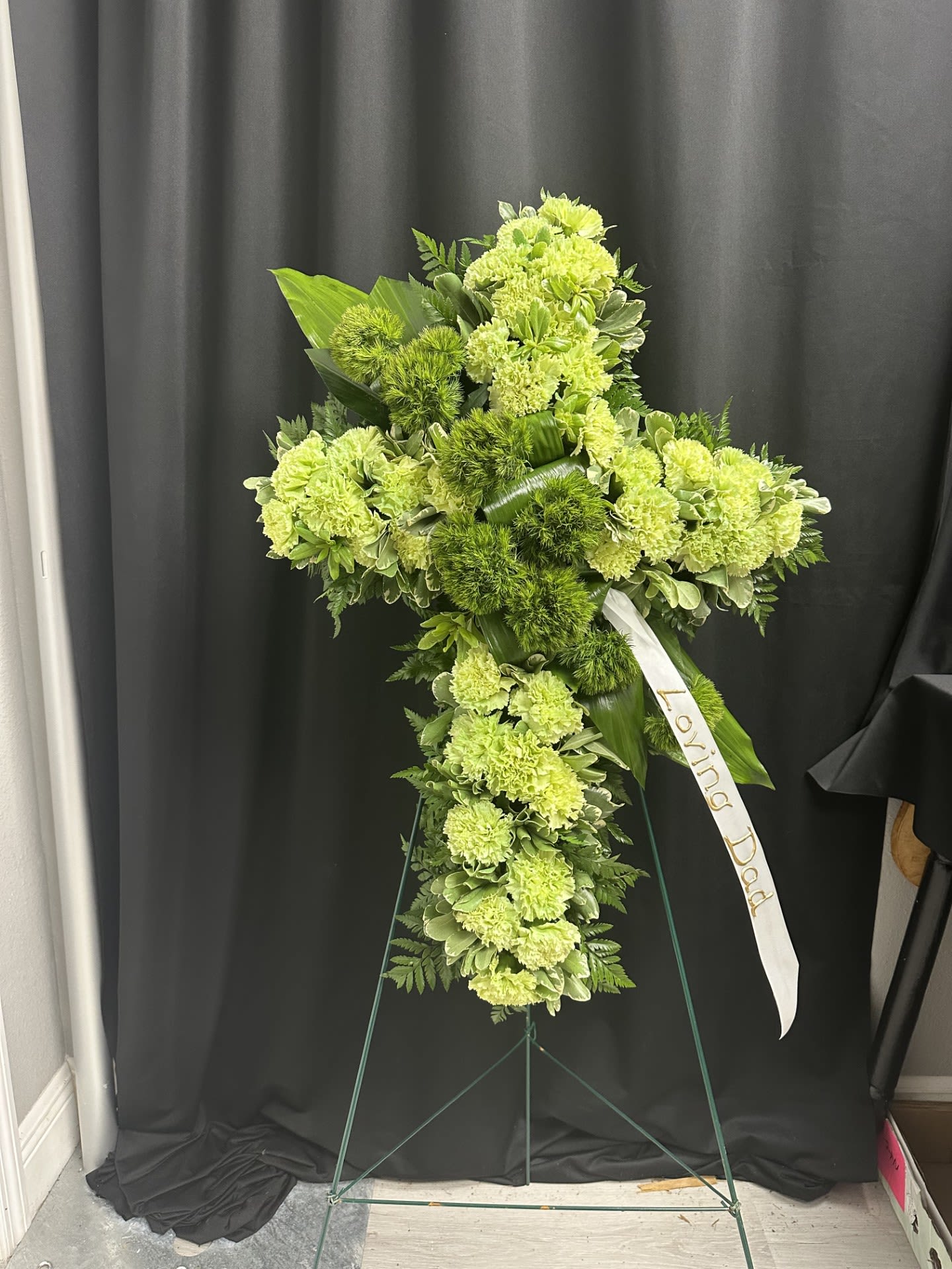 Green floral standing cross with a white ribbon on an easel