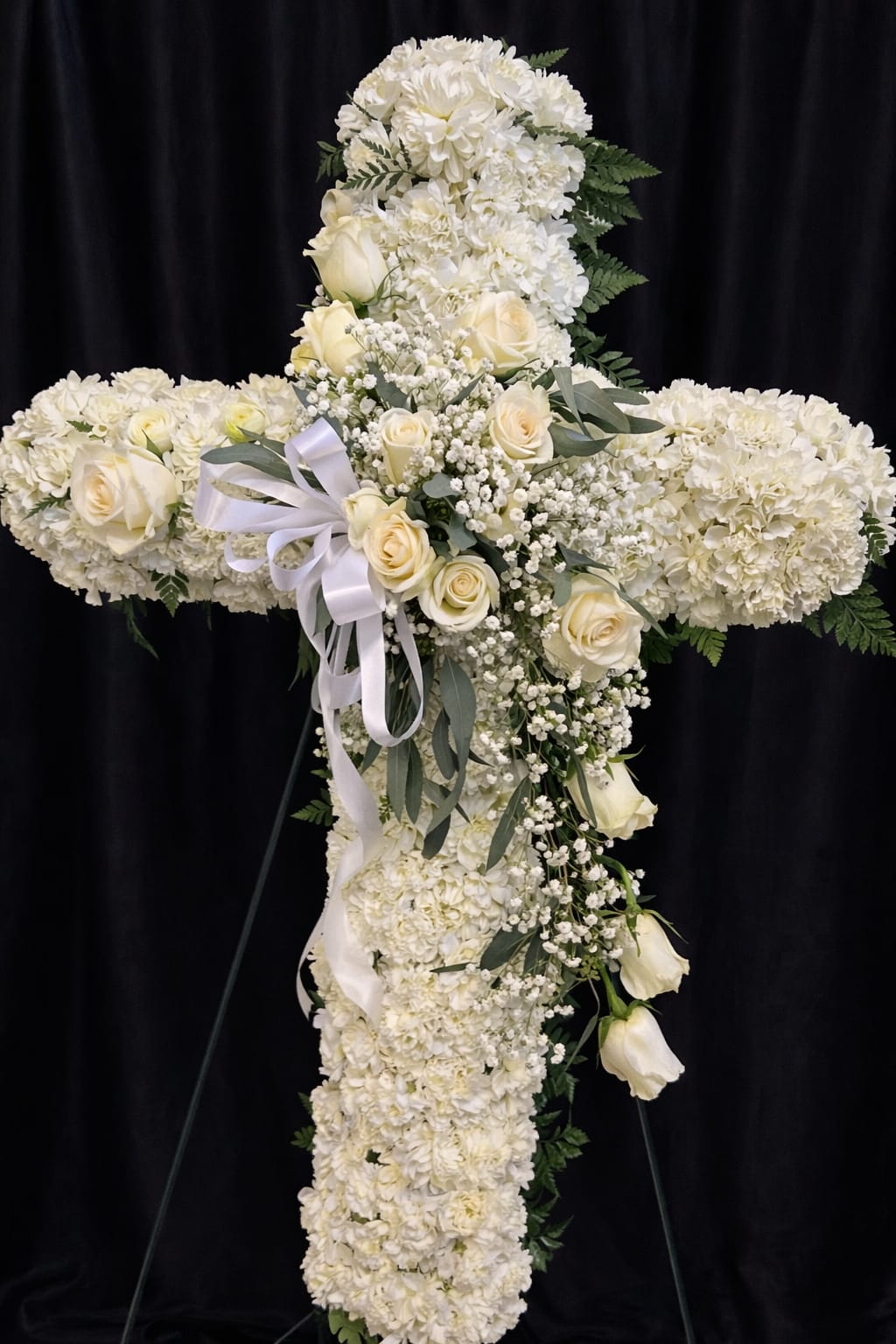 Tall white floral cross on an easel with roses and a white ribbon bow.