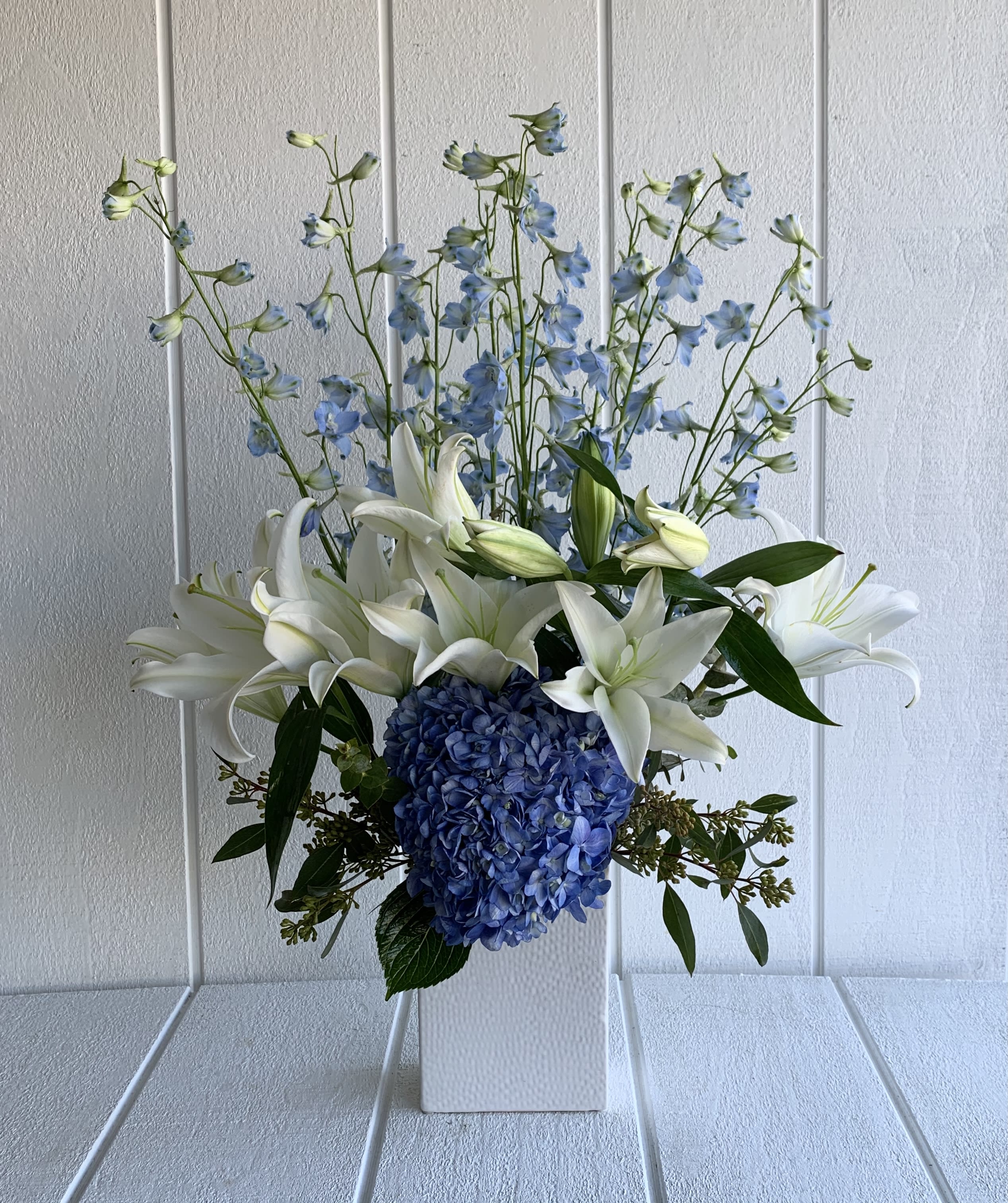 Elegant Blue - A gathering of blue and white that looks best for any special occasion. 8" H x 6" W x 3" D white ceramic vase standing at approximately 25" tall x 20" wide. The colors of Hydrangea &amp; Delphinium are subject to change depending on the market availability (dark or light blue). This picture represents deluxe size.  Availability: 24 hours notice