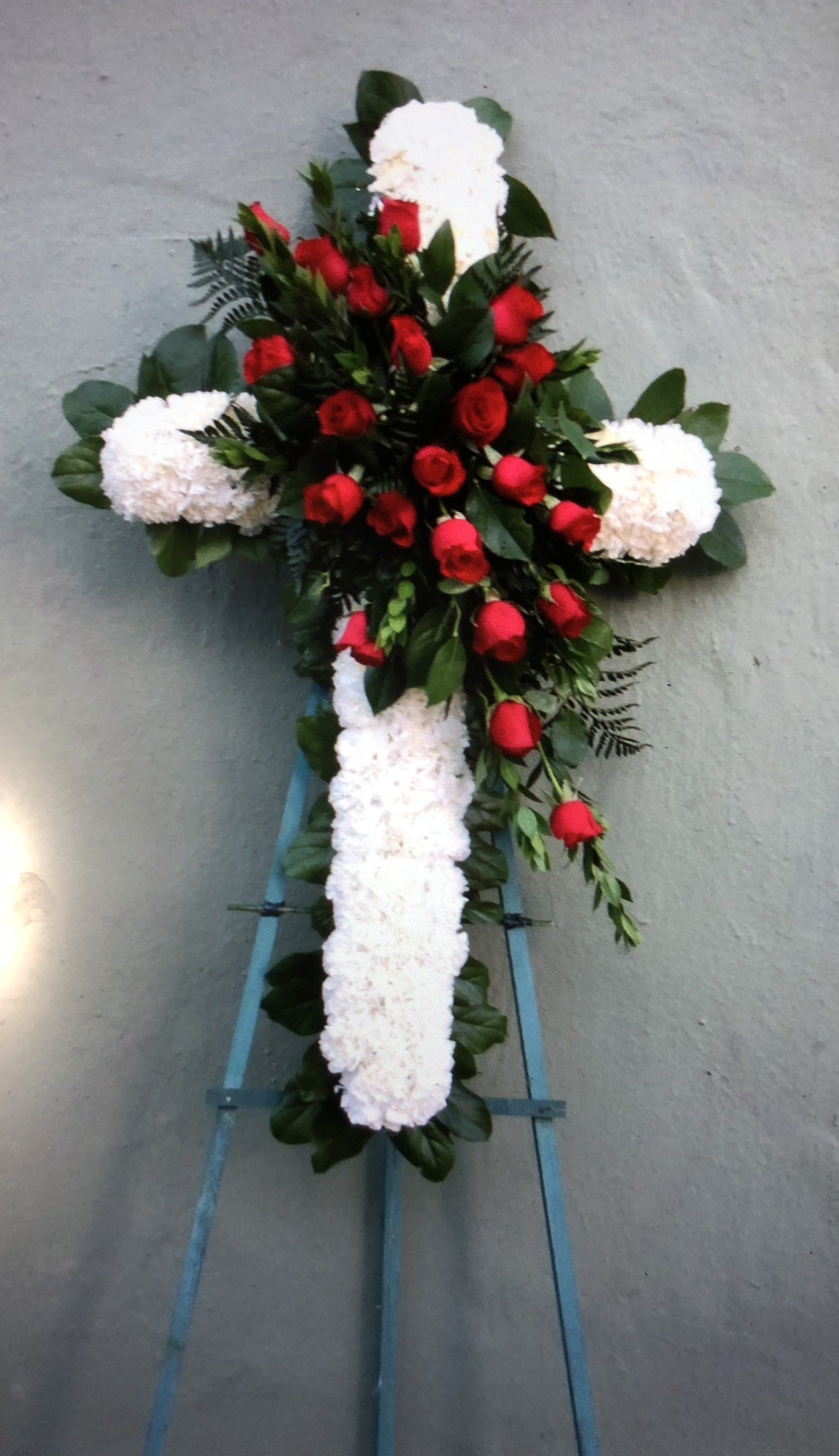 Cross-shaped floral tribute with red roses and white flowers on a stand