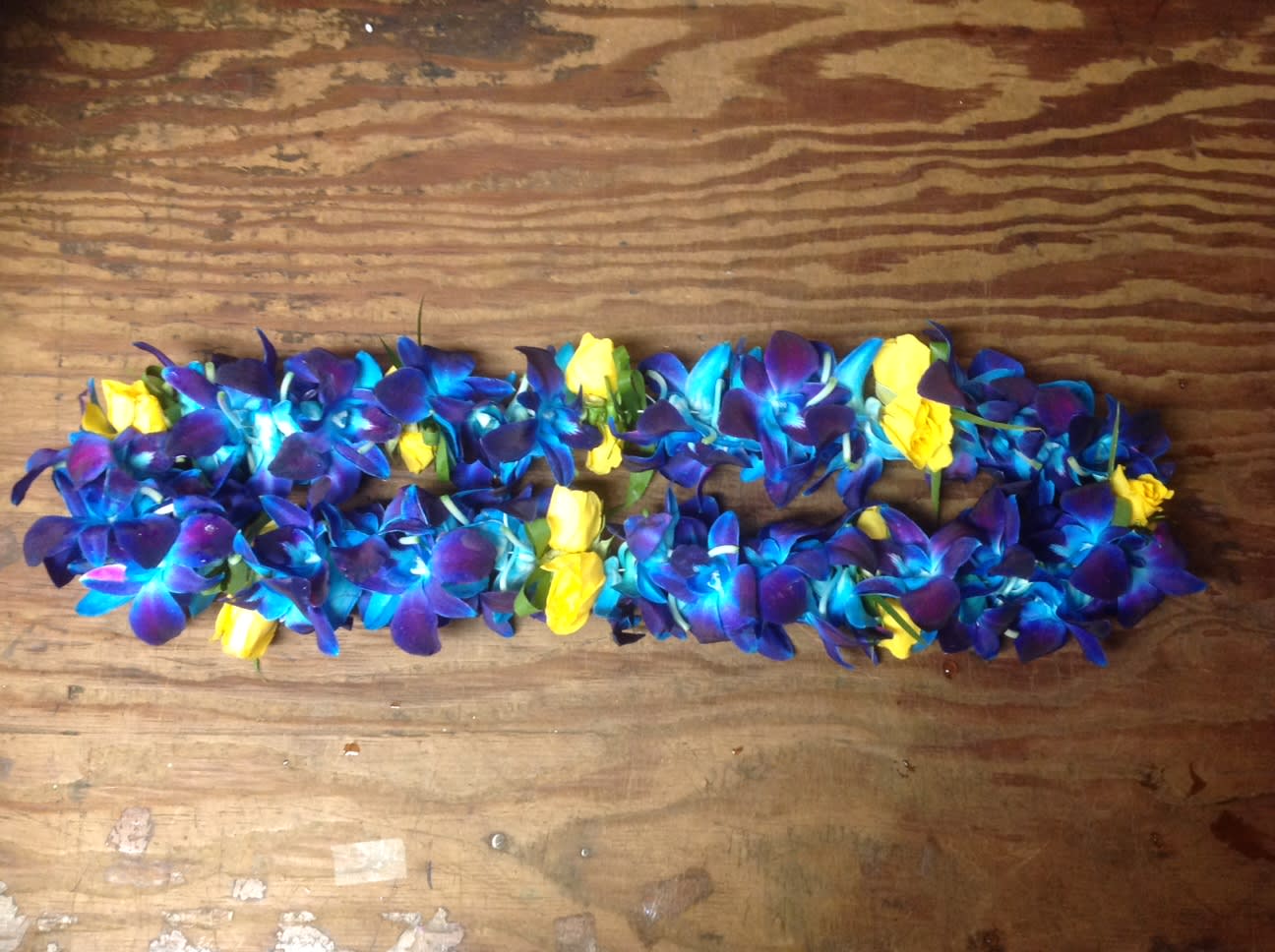 Blue and purple flower lei with yellow rose accents on a wooden surface