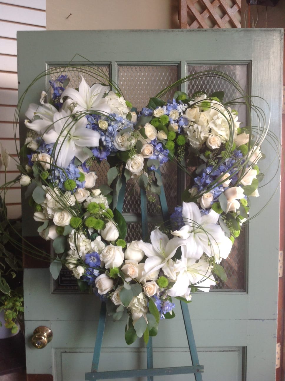 Heart-shaped floral wreath with white lilies, roses, and blue flowers on an easel