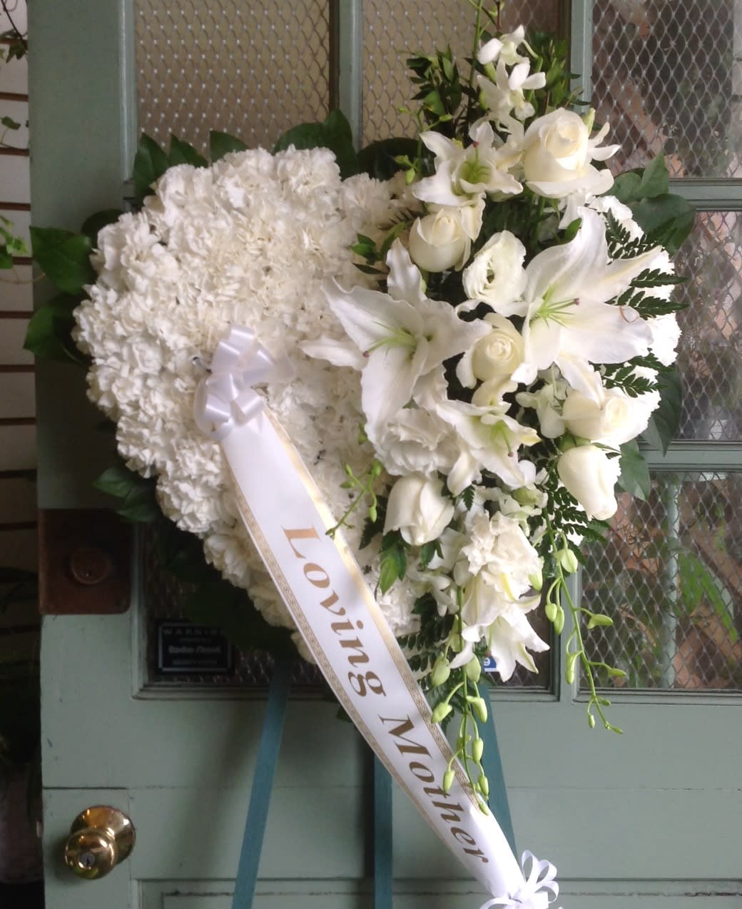 White funeral wreath with lilies and roses and a ribbon reading Loving Mother