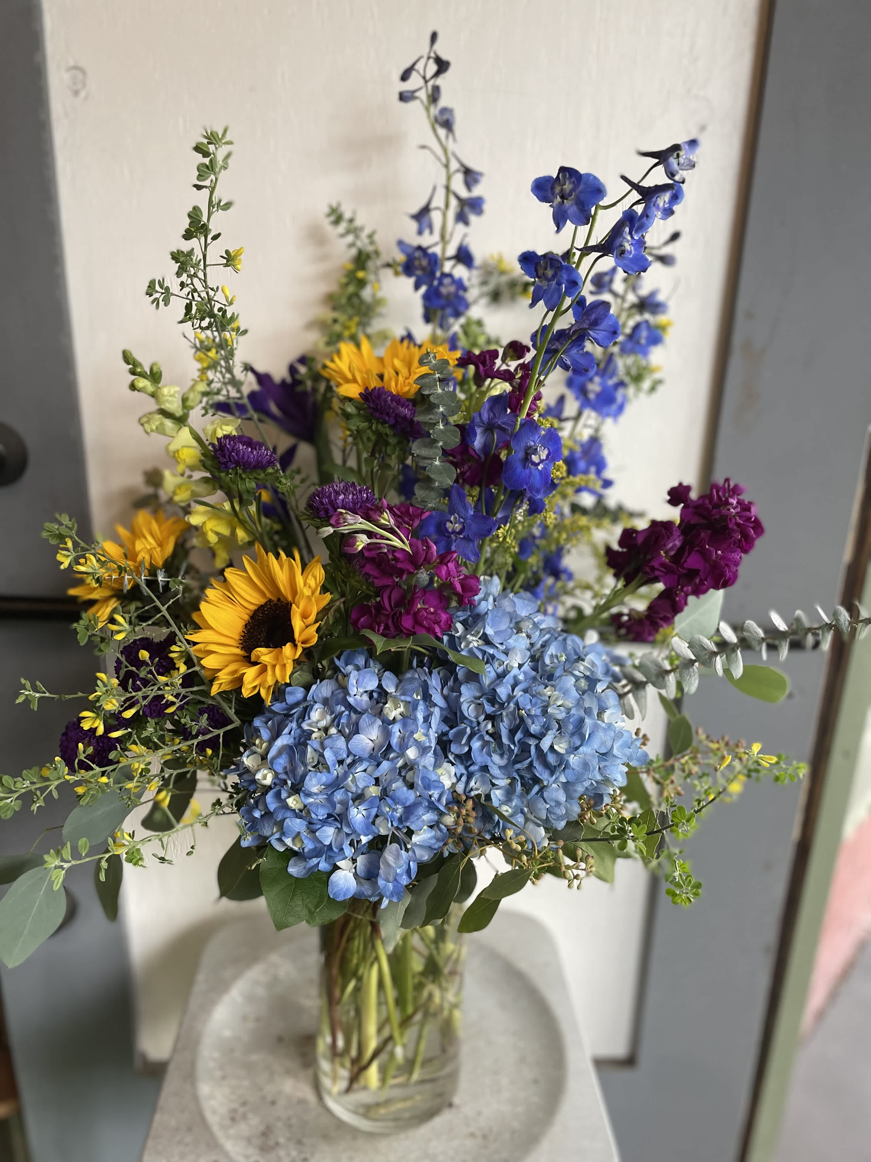 Mixed bouquet with blue hydrangeas, sunflowers, and purple blooms in a glass vase