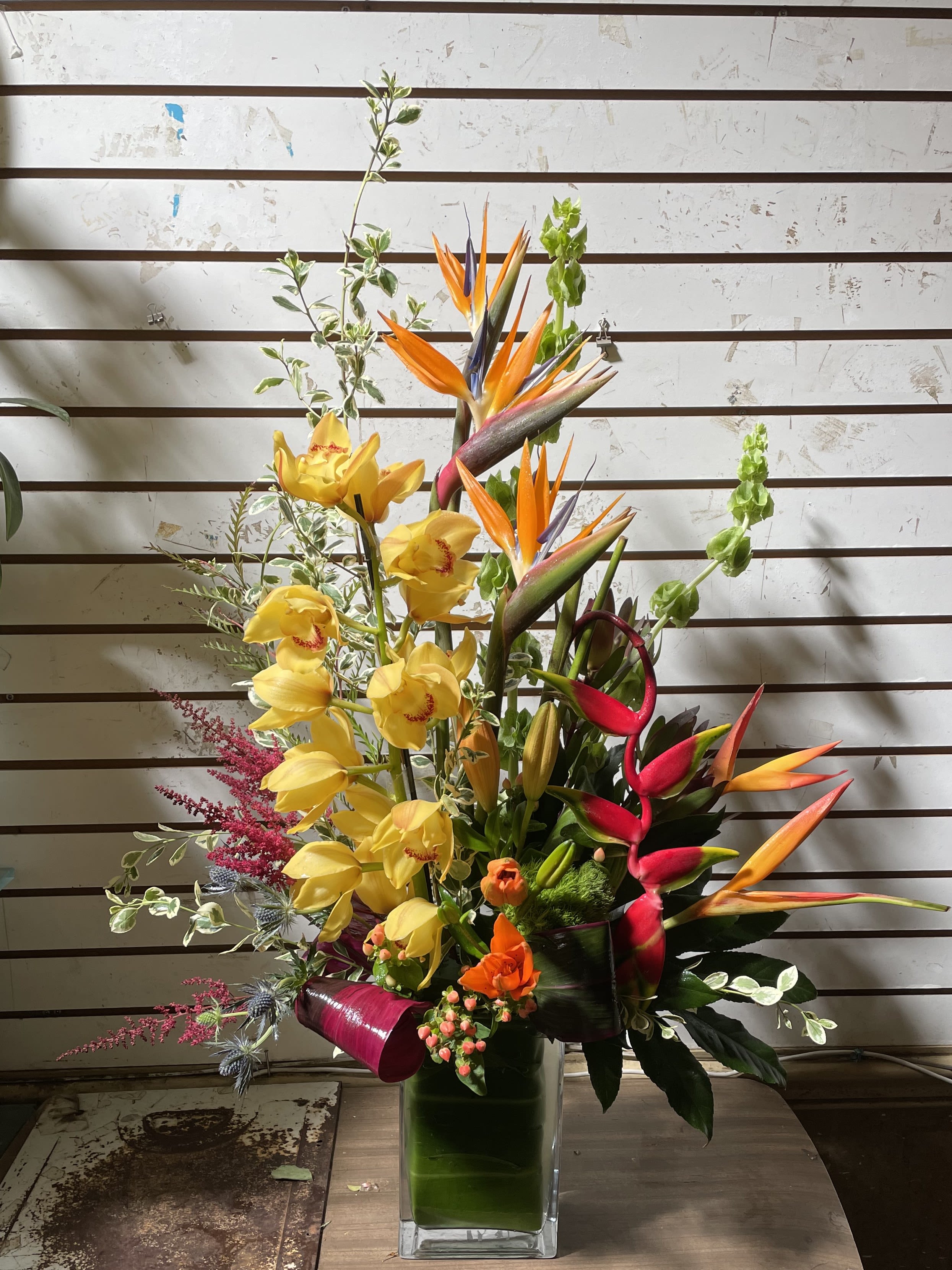 Tall tropical arrangement with yellow orchids and orange blooms in a green leaf-lined glass vase