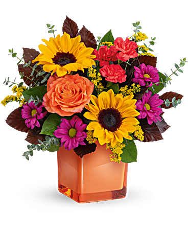 Bright mixed arrangement with sunflowers, orange rose, pink carnations and purple mums in an orange cube vase