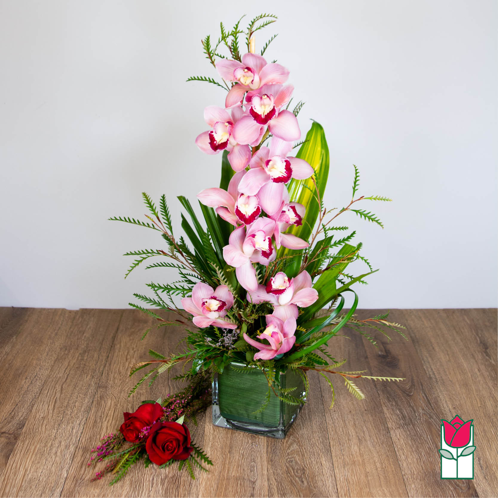 Tall arrangement of pink cymbidium orchids in a green-lined glass cube with two red roses beside it