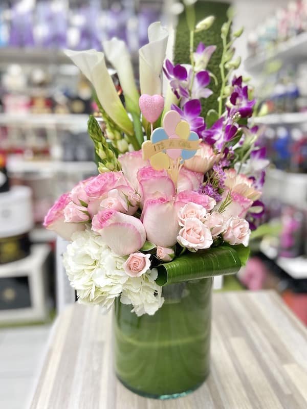 Tall arrangement of pink and white flowers with calla lilies in a green glass vase and an 'It's a Girl' pick